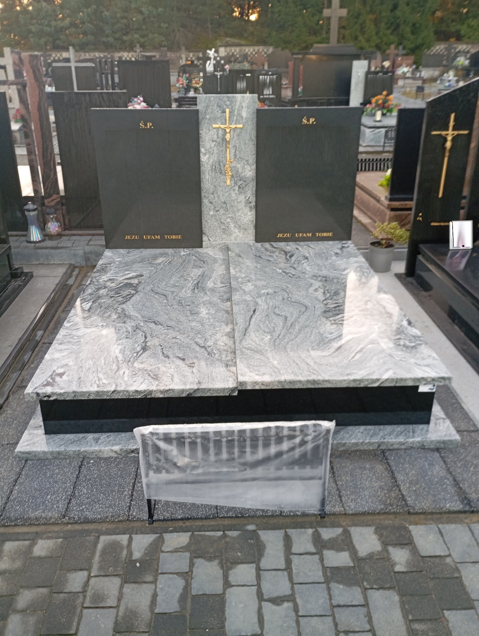 Granitowy nagrobek z szarą płytą w faliste wzory, złotym krzyżem i napisem 'Jezu ufam Tobie', na cmentarzu z kostką brukową i innymi nagrobkami w tle.