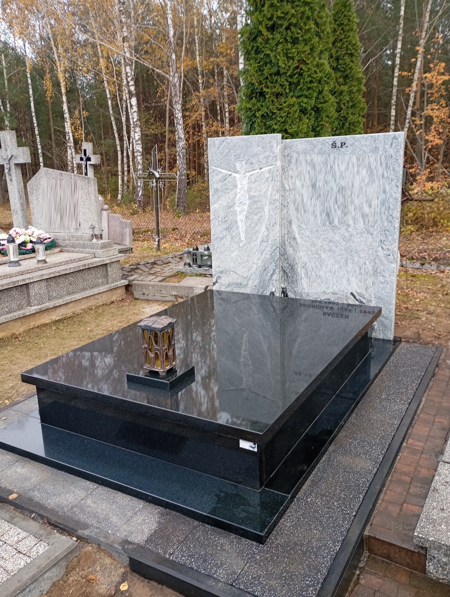 Czarny, polerowany nagrobek z szarego granitu z wizerunkiem Chrystusa, otoczony jesiennym krajobrazem cmentarza. Na płycie nagrobnej stoi lampion.