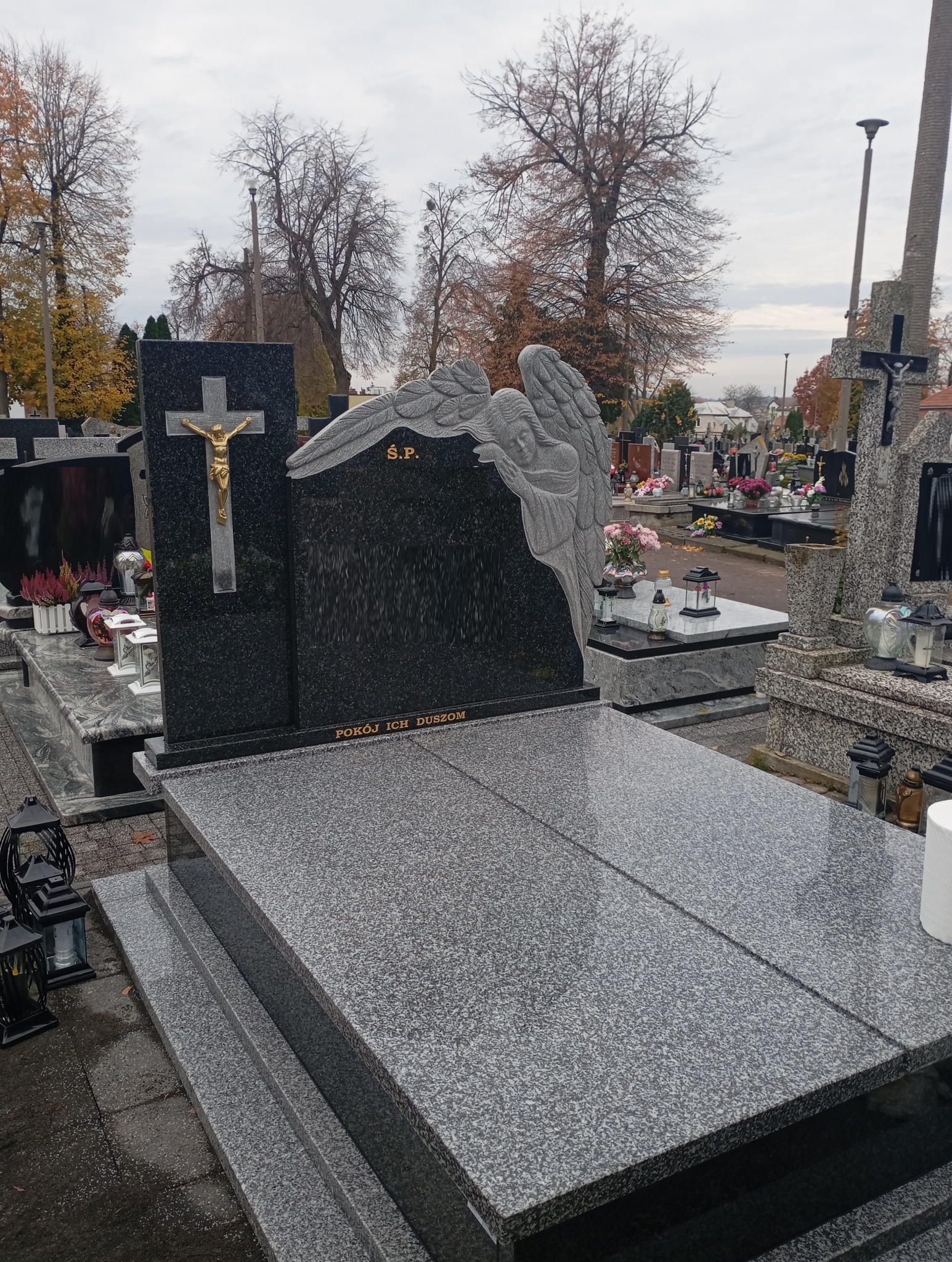 Granitowy nagrobek z rzeźbą anioła i krzyżem na cmentarzu, jesienna sceneria. Napis 'S.P. Pokój ich dusz'.