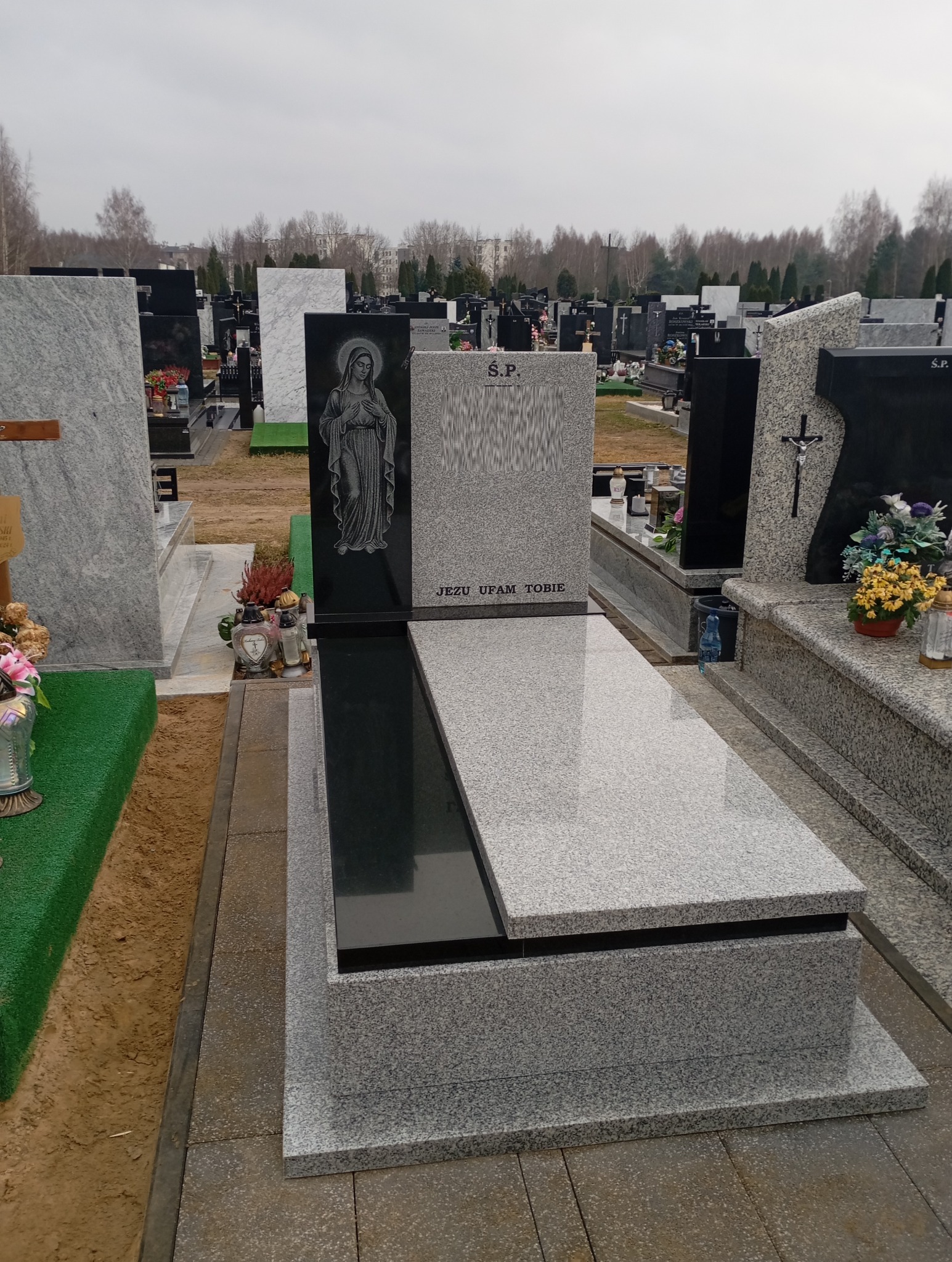 Granitowy nagrobek z wizerunkiem Matki Boskiej i napisem 'Jezu ufam Tobie' na cmentarzu, w tle inne nagrobki i drzewa. Szara kolorystyka, pochmurne niebo.