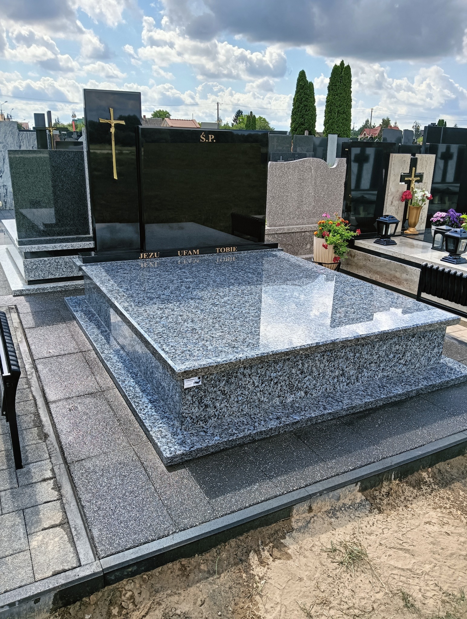 Granitowy nagrobek z inskrypcją 'Jezu ufam Tobie' i złotym krzyżem na czarnej płycie, otoczony innymi nagrobkami na cmentarzu w słoneczny dzień.
