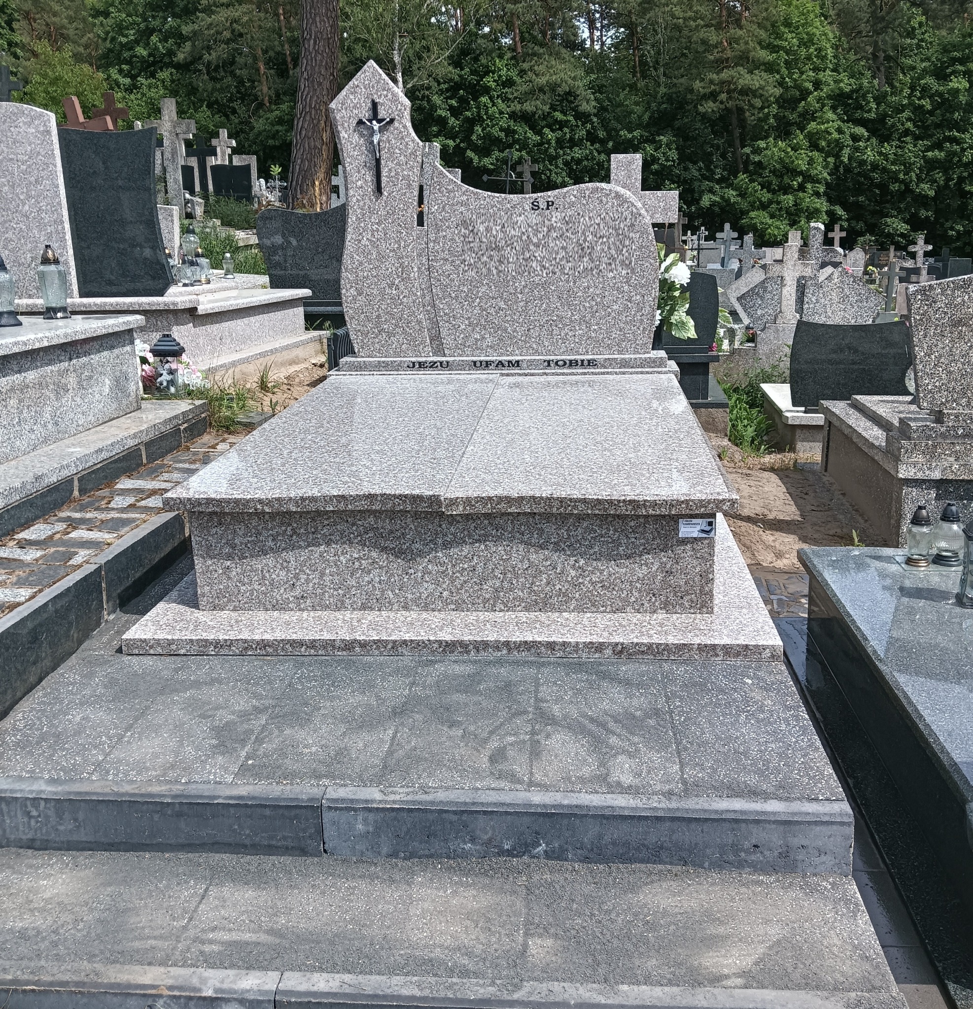 Jasnoszary, granitowy nagrobek z krzyżem i napisem 'Jezu ufam Tobie' na cmentarzu, otoczony zielenią drzew. Widok z przodu, w tle inne nagrobki.