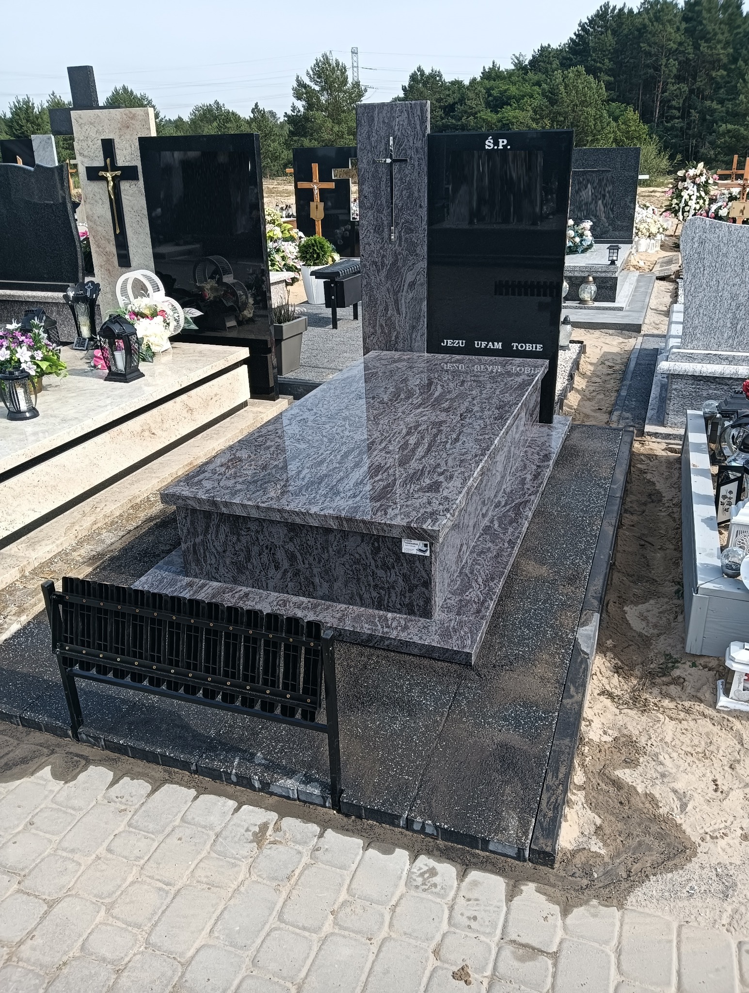 Granitowy nagrobek z napisem 'Jezu ufam Tobie' na cmentarzu, otoczony innymi nagrobkami i zielenią. Widoczna metalowa barierka i kostka brukowa.