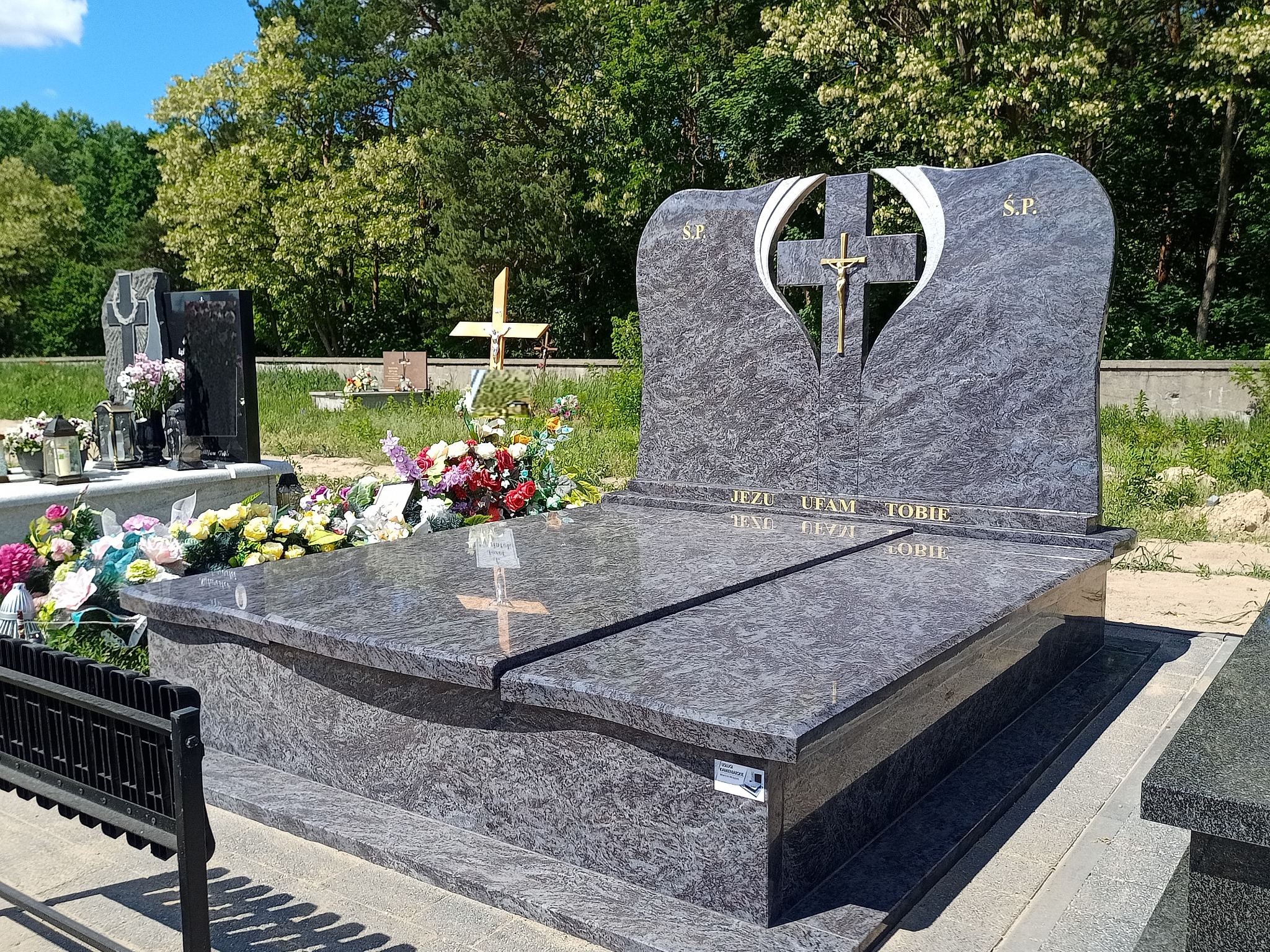 Granitowy nagrobek z krzyżem i napisem 'Jezu ufam Tobie' na tle zieleni cmentarza.