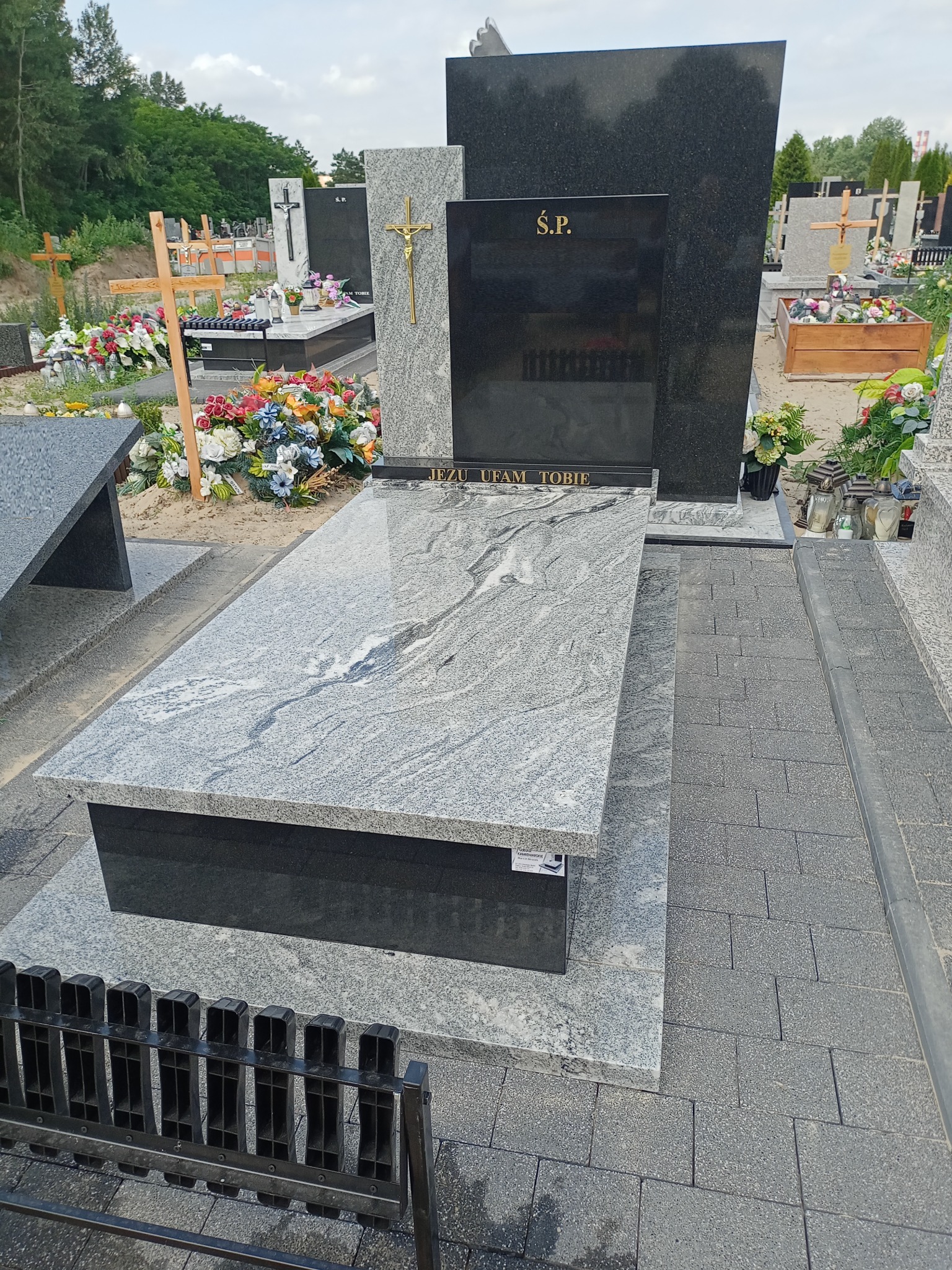 Nowoczesny nagrobek z szarego i czarnego granitu, z napisem 'Jezu ufam Tobie' i złotym krzyżem, otoczony kostką brukową na cmentarzu.
