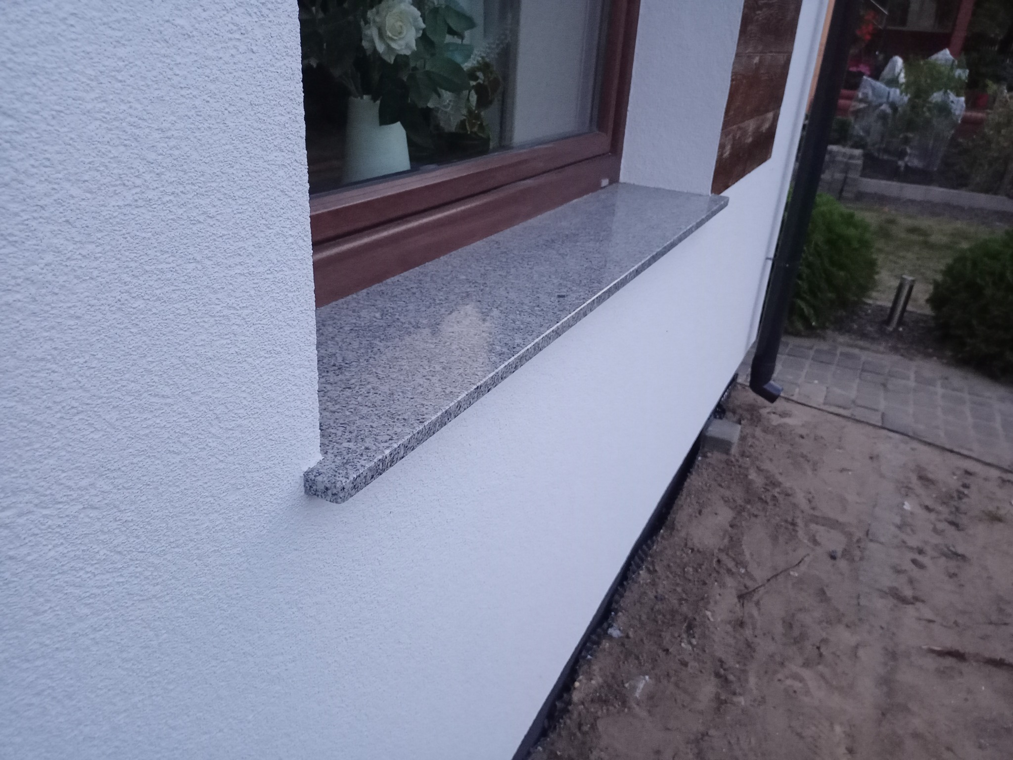 Zamontowany szary parapet zewnętrzny z konglomeratu, z zaokrąglonym brzegiem, pod oknem z brązową ramą, na tle białego, chropowatego tynku i zieleni ogrodu.