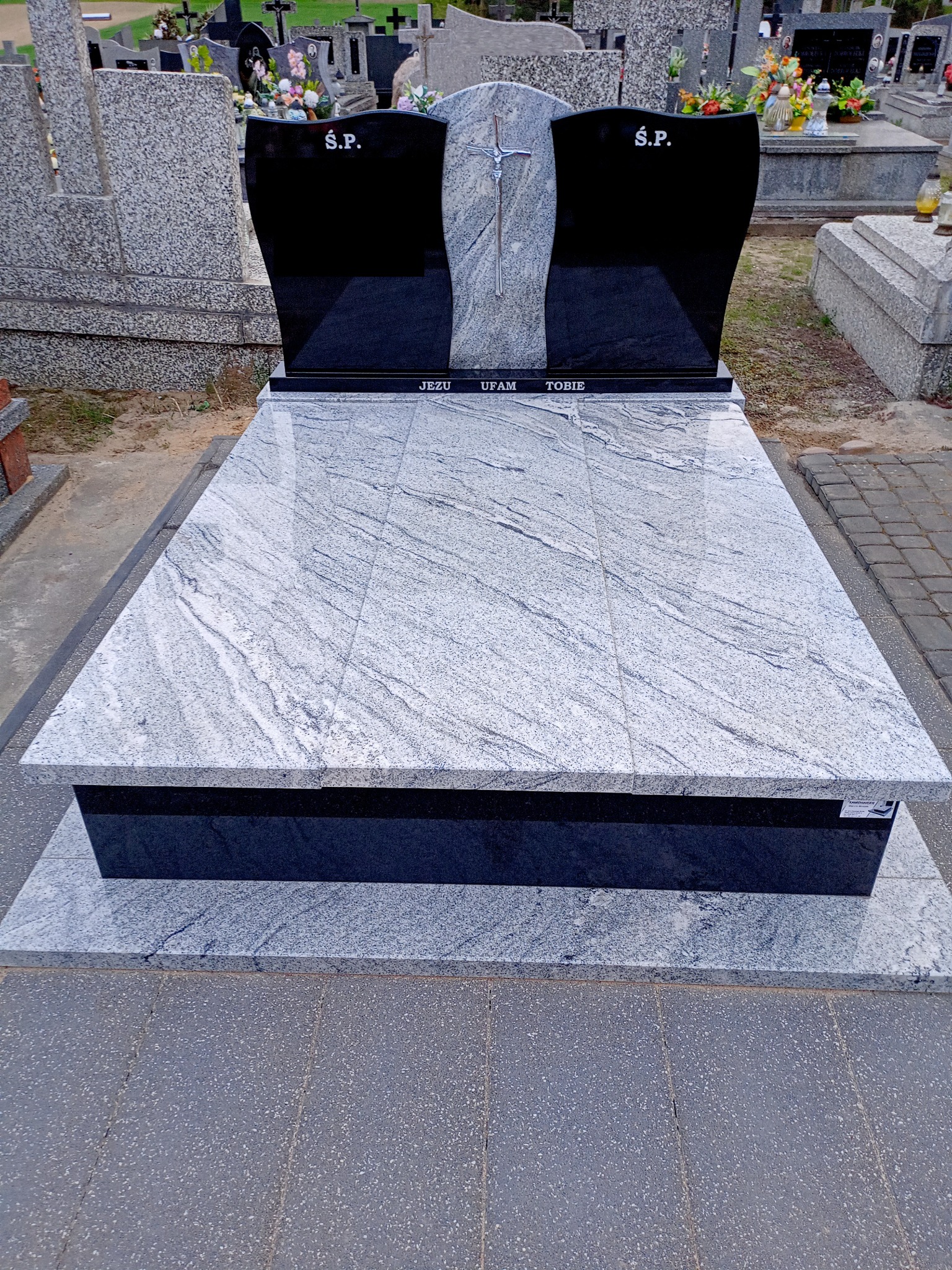 Nowoczesny, dwuczęściowy pomnik z jasnoszarego granitu z czarnymi tablicami nagrobnymi po bokach, krzyż z metalu w centralnej części, napis 'Jezu ufam Tobie' na dolnej płycie, otoczony innymi...