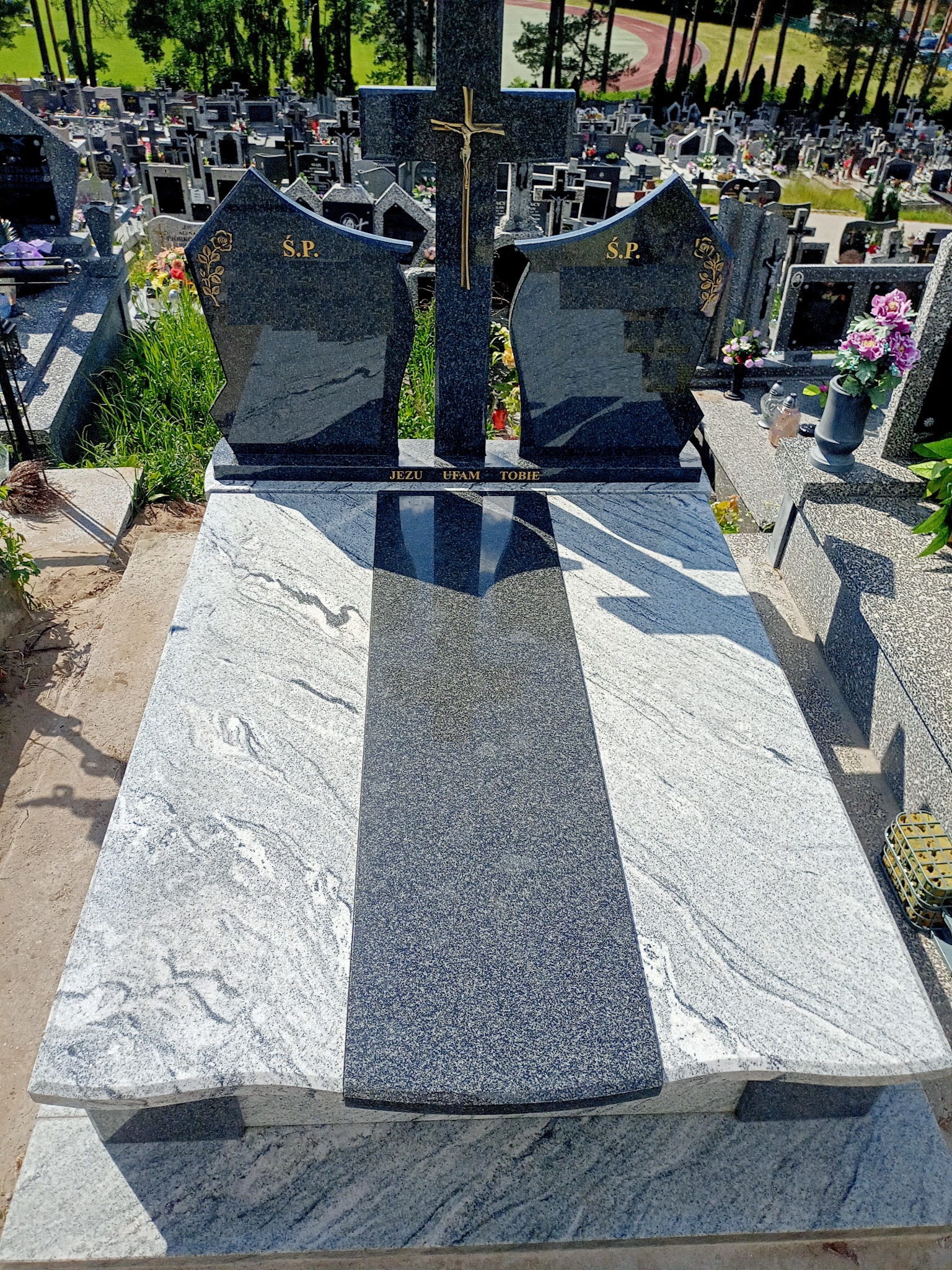 Nowoczesny nagrobek z jasnoszarego i ciemnoszarego granitu z napisem 'Jezu Ufam Tobie' na tle cmentarza.