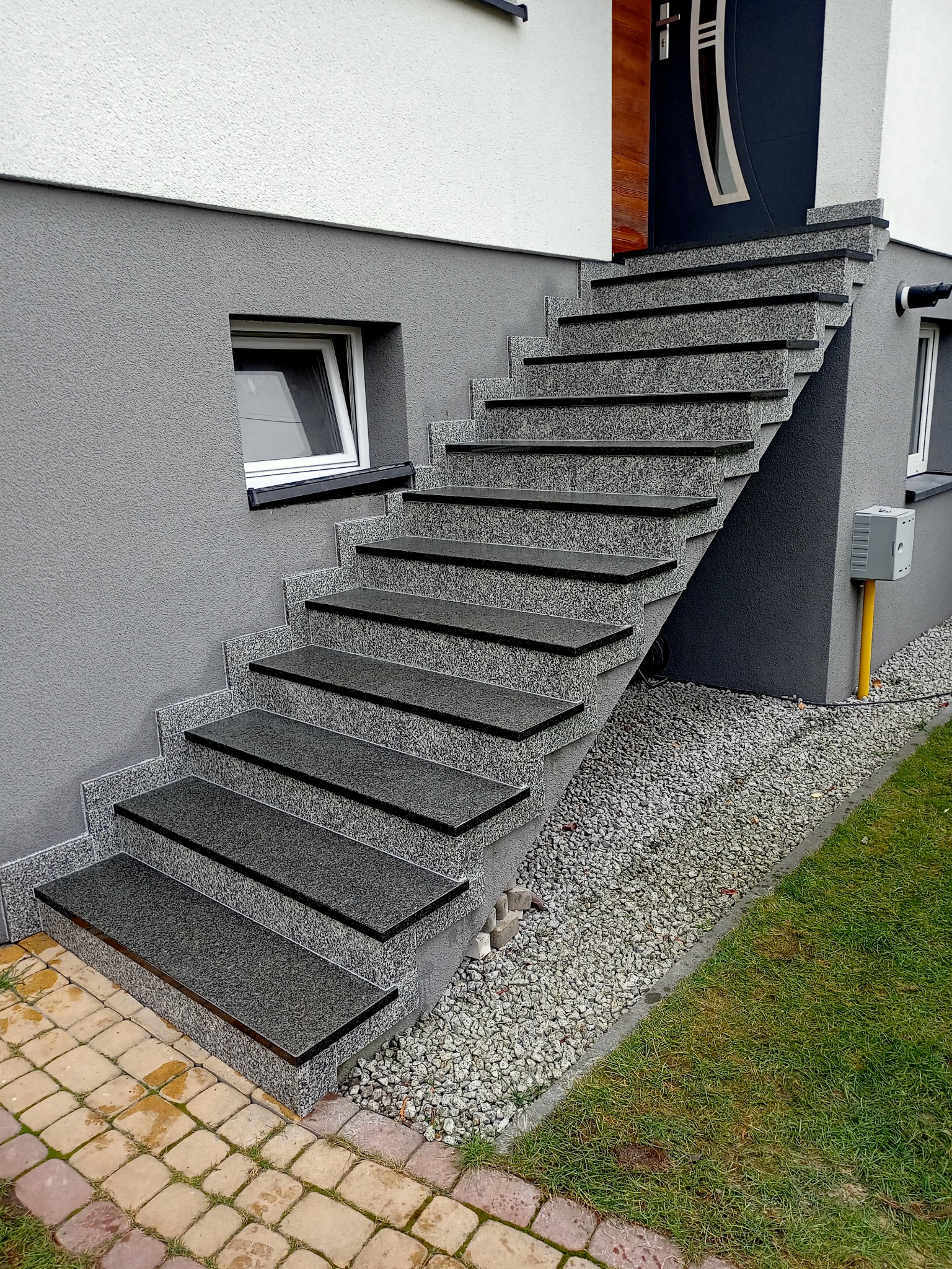 Zewnętrzne schody wejściowe z szarego kamienia z czarnymi krawędziami, prowadzące do ciemnych drzwi wejściowych w nowoczesnym domu z szarą elewacją i białym pasem na górze, otoczone jasnym żwirem...