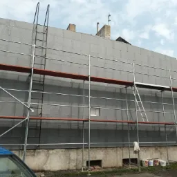 Budynek w trakcie ocieplania, widoczne szare płyty izolacyjne na elewacji, rusztowanie, drabina oparta o ścianę, oraz dekoracja ogrodowa w kształcie wiedźmy na miotle.