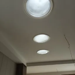 Trzy okrągłe świetliki w białym suficie podwieszanym, widoczne fragmenty zabudowy meblowej i okna z żaluzjami.