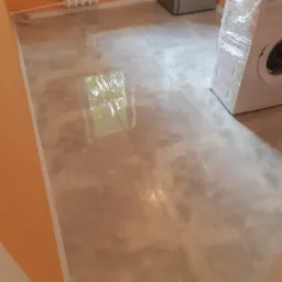 Beżowe płytki podłogowe imitujące kamień, świeżo położone w łazience, z widoczną pralką owiniętą folią ochronną i fragmentem grzejnika na pomarańczowej ścianie