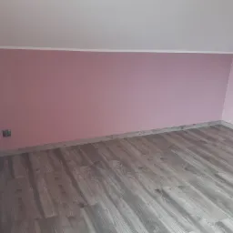 Pokój po odświeżeniu: ściany pomalowane na delikatny róż, podłoga wyłożona panelami w odcieniach szarości imitujących drewno, biała listwa przypodłogowa i grzejnik.