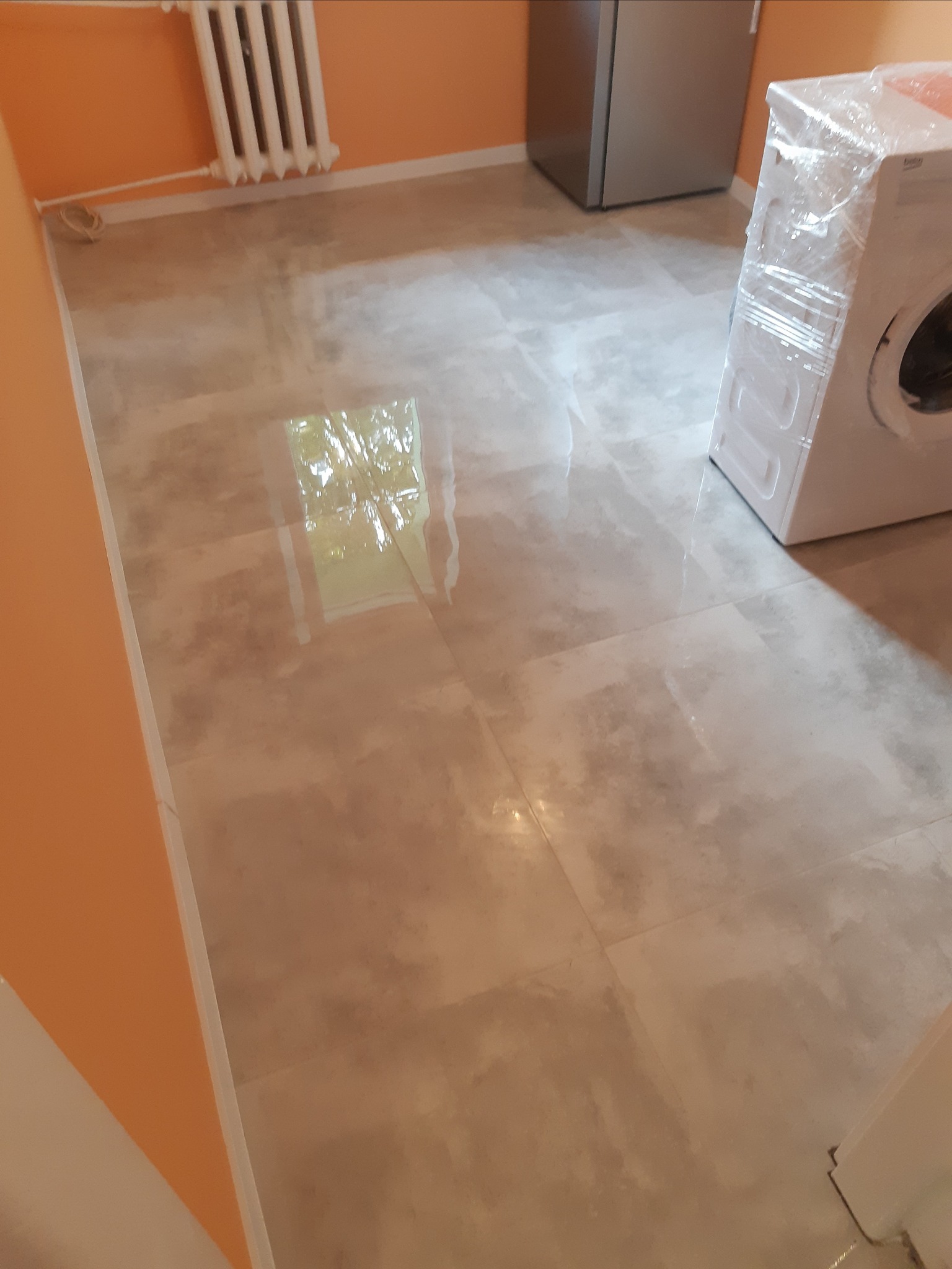 Beżowe płytki podłogowe imitujące kamień, świeżo położone w łazience, z widoczną pralką owiniętą folią ochronną i fragmentem grzejnika na pomarańczowej ścianie