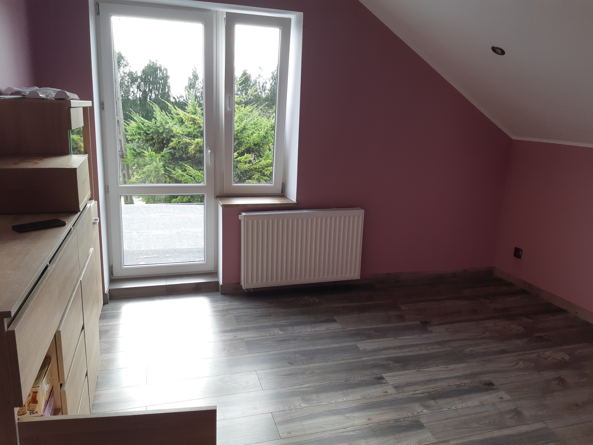 Poddasze z nową podłogą imitującą drewno, pomalowane ściany na różowo, biały grzejnik pod oknem z widokiem na zieleń.