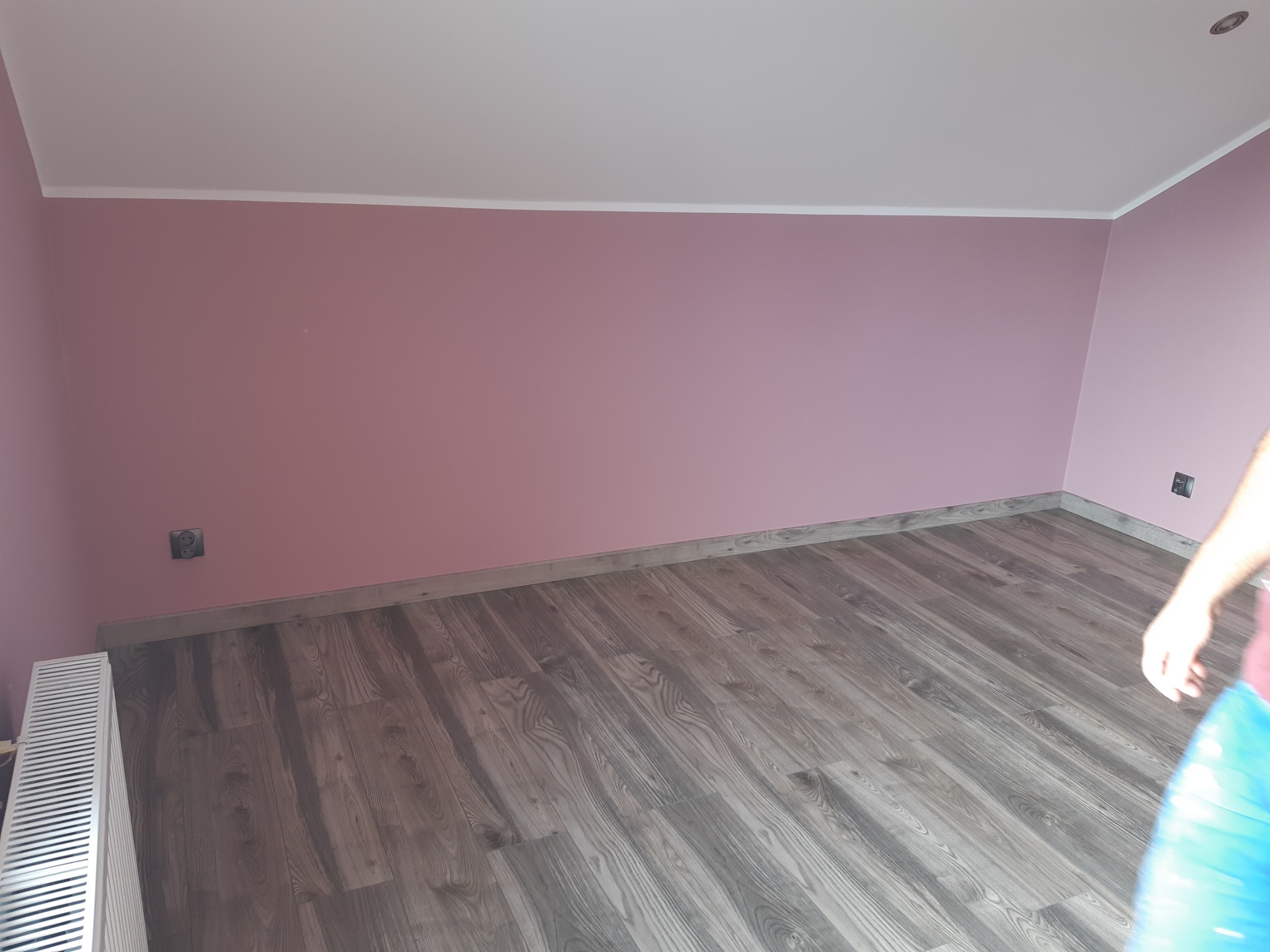 Pokój po odświeżeniu: ściany pomalowane na delikatny róż, podłoga wyłożona panelami w odcieniach szarości imitujących drewno, biała listwa przypodłogowa i grzejnik.