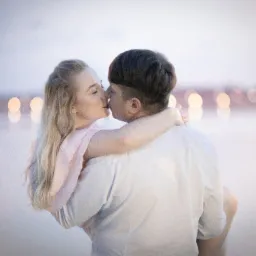 Narzeczona w różowej sukience, trzymana w ramionach przez narzeczonego, pocałunek na tle rozmytych świateł miasta.