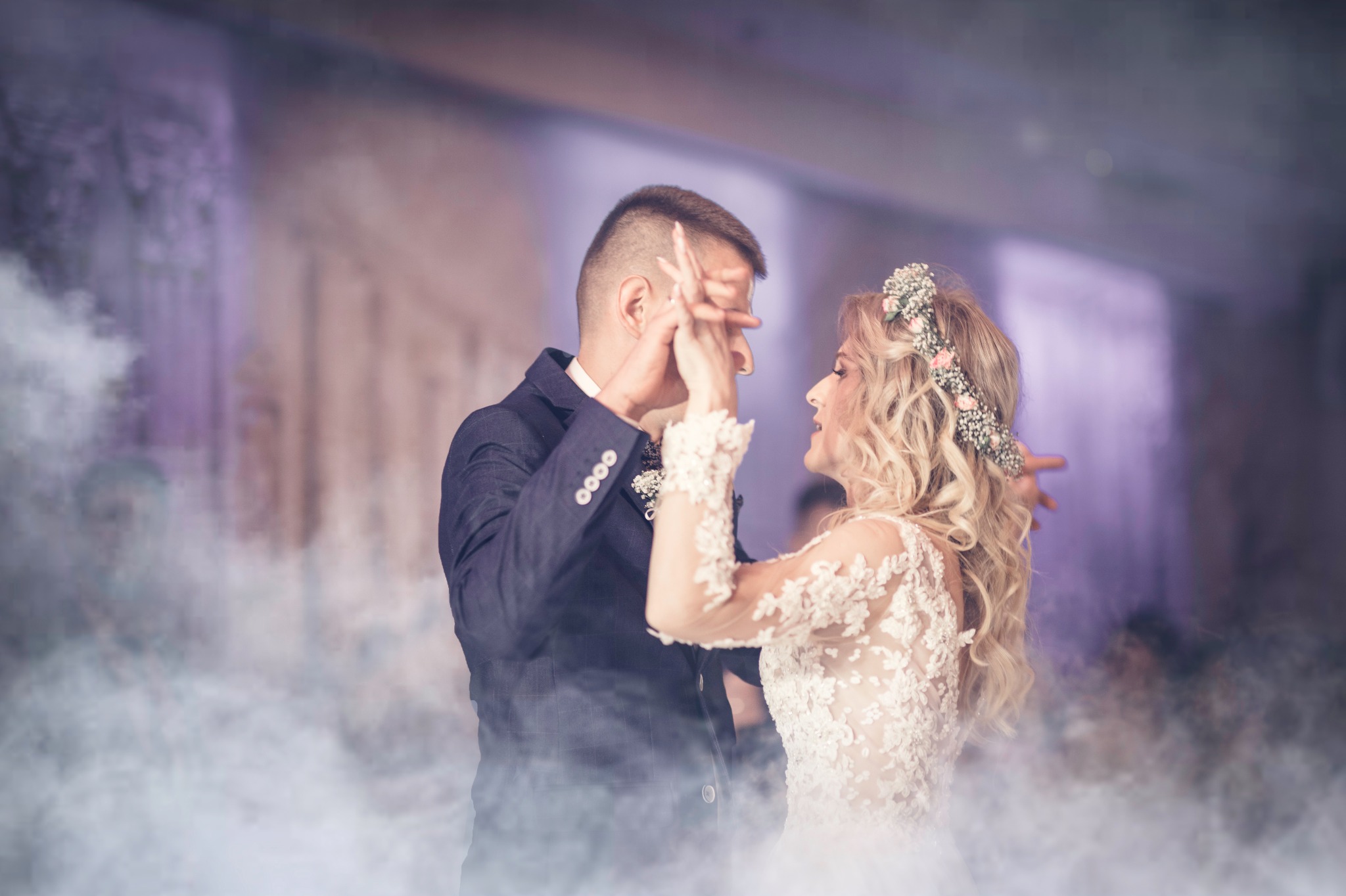 Para młoda w tańcu, panna młoda w koronkowej sukni i wianku, pan młody w granatowym garniturze, otoczeni mgłą na sali balowej.