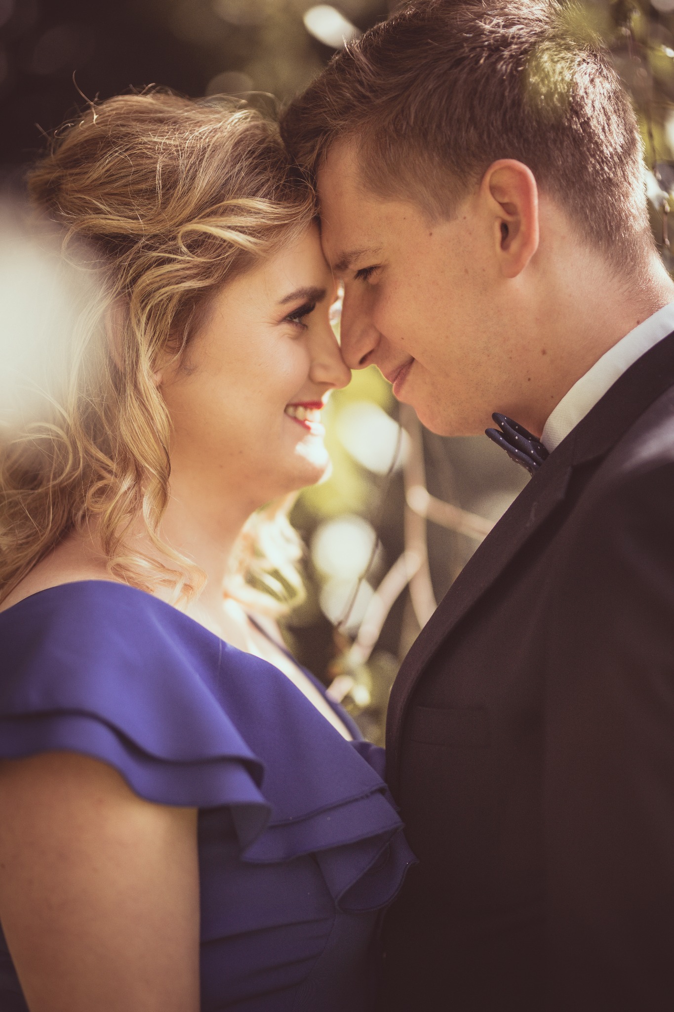 Uśmiechnięta para, zbliżone twarze, ona w kobaltowej sukience z falbanami, on w ciemnym garniturze z muszką w kropki, delikatne oświetlenie.