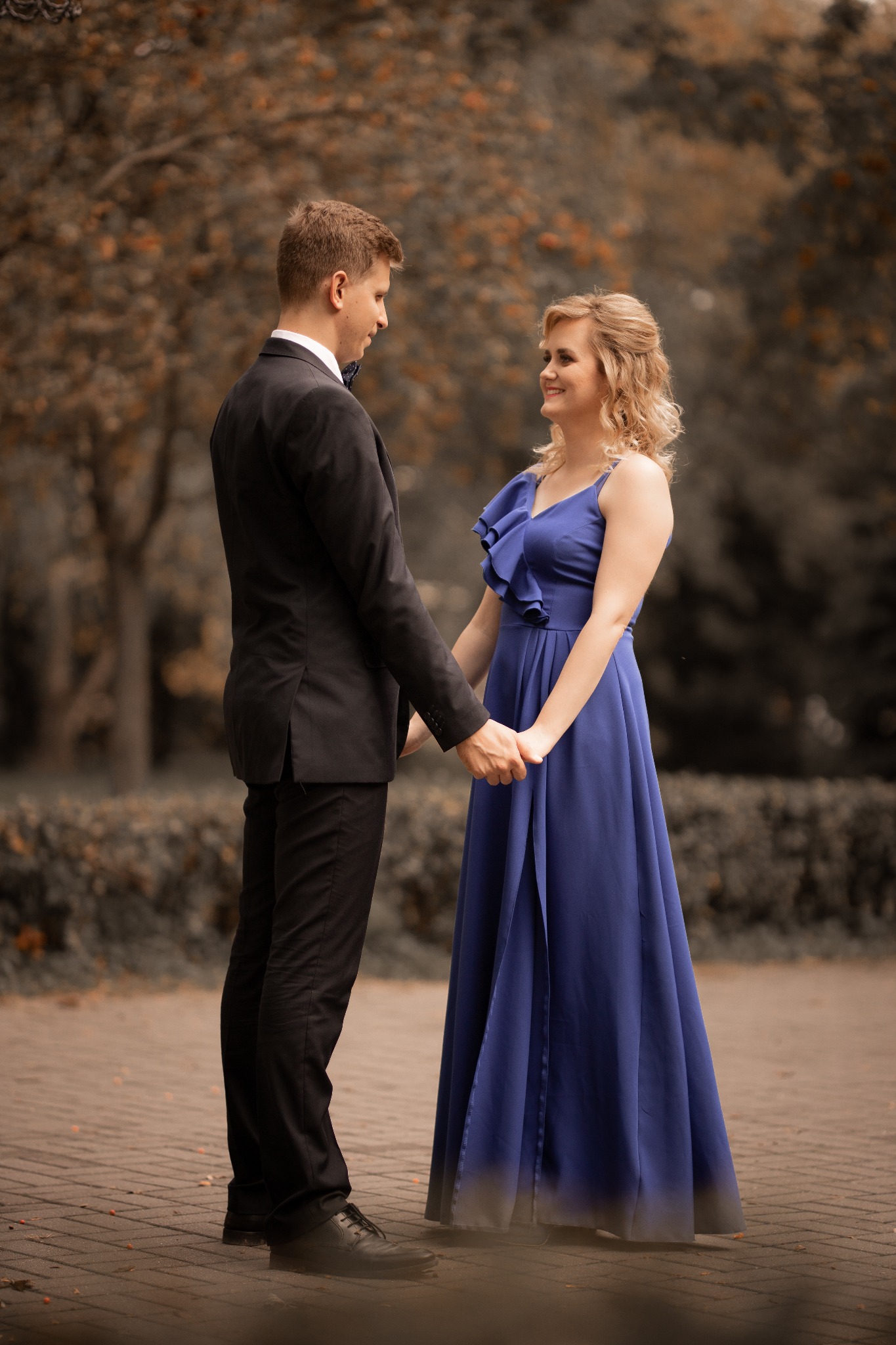 Para trzymająca się za ręce, on w czarnym garniturze z muchą, ona w długiej, kobaltowej sukni z falbanami, oboje patrzą na siebie z uśmiechem, jesienne tło z drzewami.