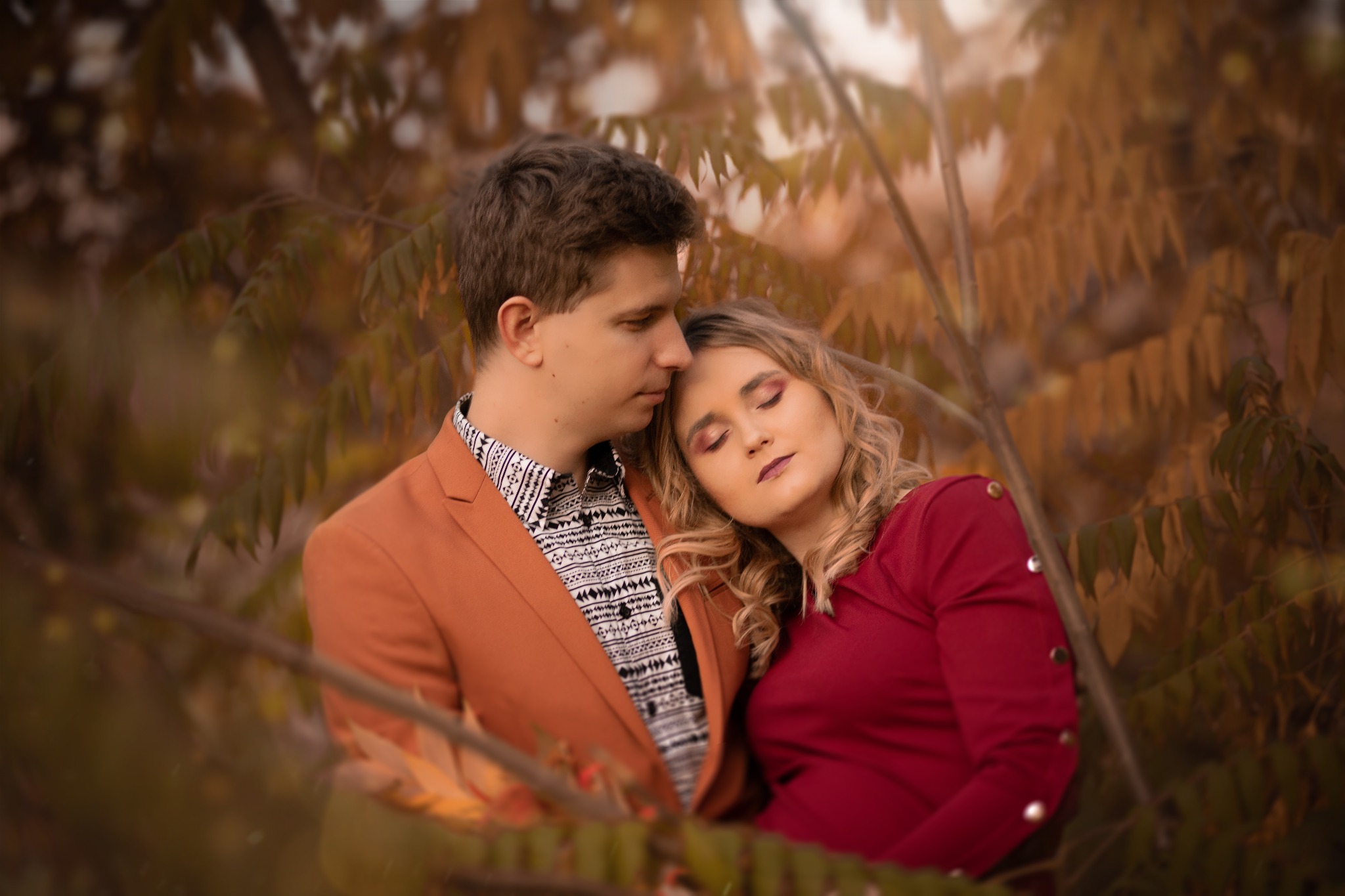 Para zakochanych w jesiennej scenerii: mężczyzna w brązowej marynarce i wzorzystej koszuli przytula kobietę w czerwonej sukience na tle drzew z opadającymi liśćmi.