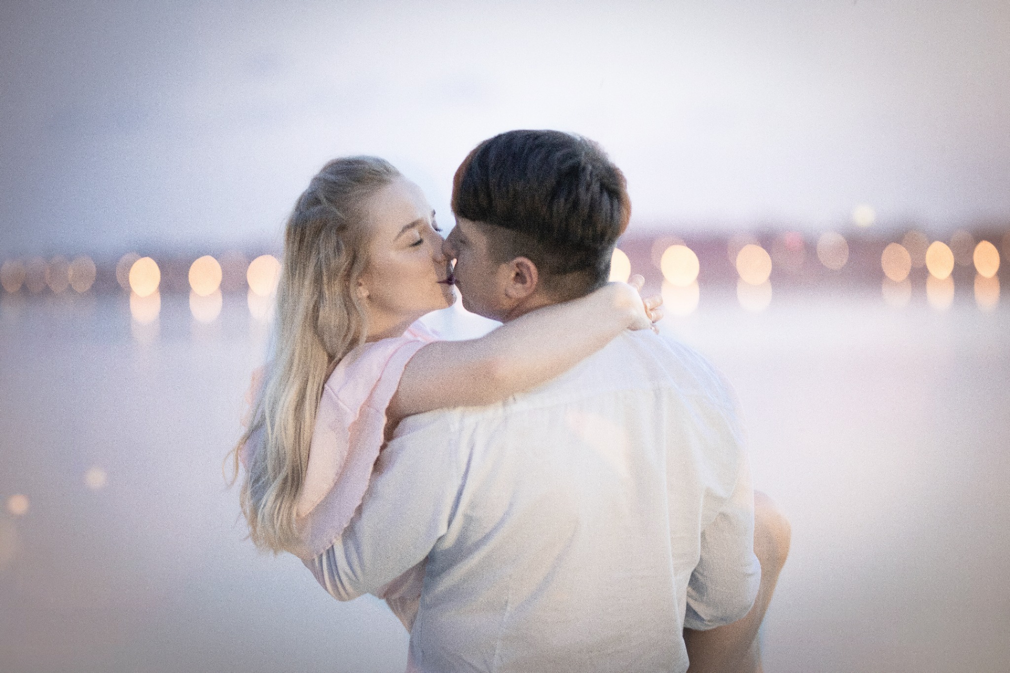 Narzeczona w różowej sukience, trzymana w ramionach przez narzeczonego, pocałunek na tle rozmytych świateł miasta.