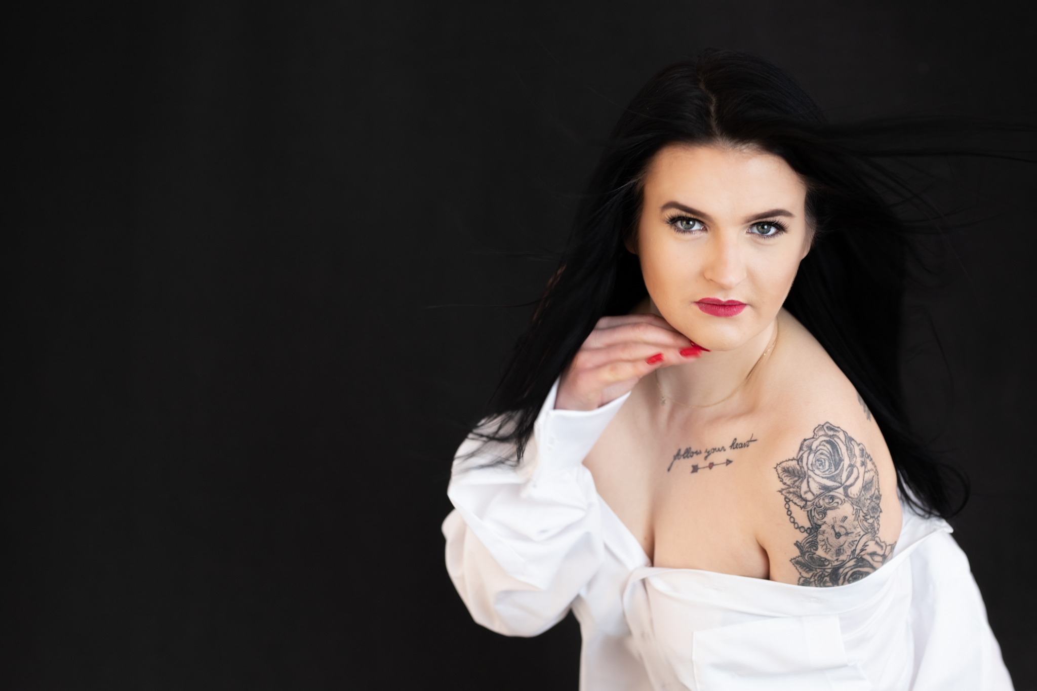 Portret kobiety z tatuażem róży i zegara na ramieniu, w białej koszuli na czarnym tle, z wiatrem we włosach.