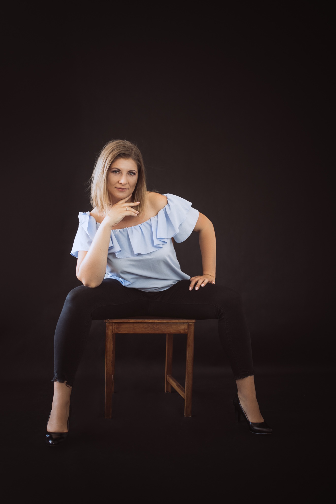Portret kobiety w niebieskiej bluzce i czarnych spodniach, siedzącej na drewnianym krześle na ciemnym tle, wykonany w Lublinie.