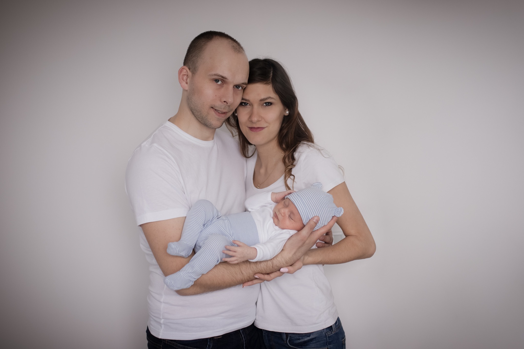 Rodzinne ujęcie: młoda para w białych koszulkach trzyma na rękach śpiącego noworodka w pasiastym ubranku i czapeczce z uszkami na tle jasnej ściany.