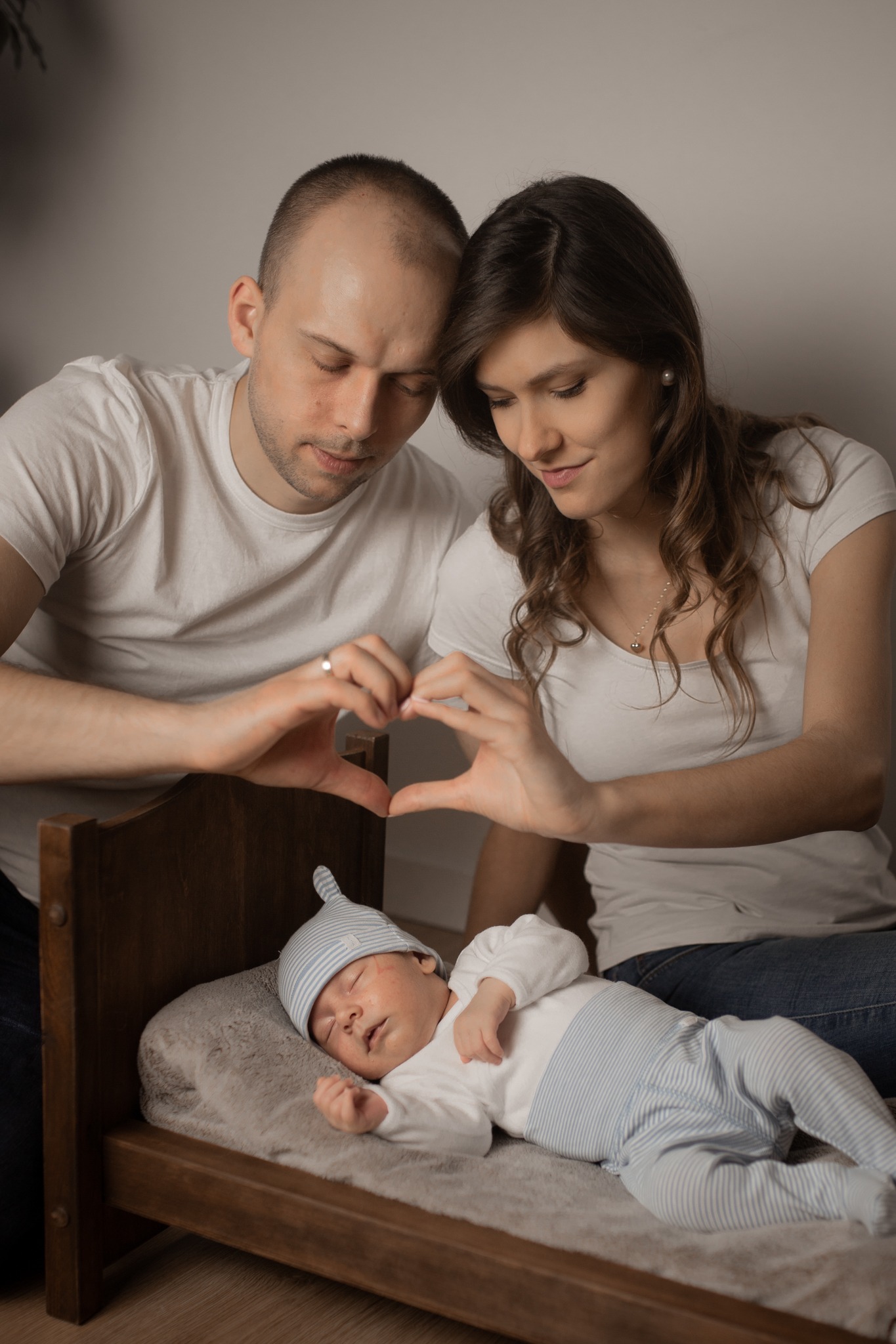 Rodzice tworzą dłońmi kształt serca nad śpiącym noworodkiem w pasiastym ubranku i czapce, leżącym w drewnianym łóżeczku.