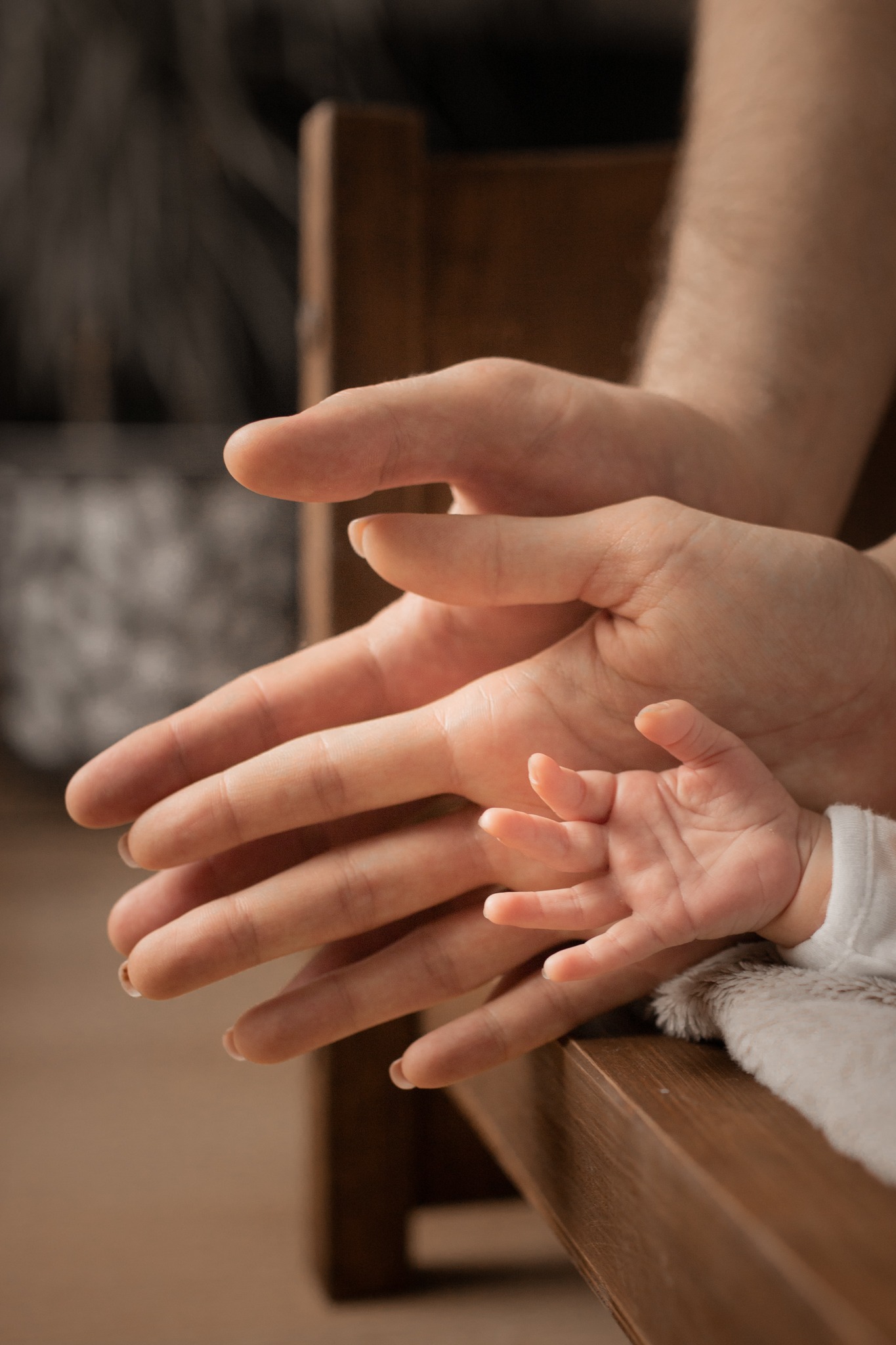 Dłoń noworodka spoczywająca na dłoniach rodziców, ułożonych na drewnianym łóżeczku, delikatne oświetlenie podkreśla więź rodzinną.