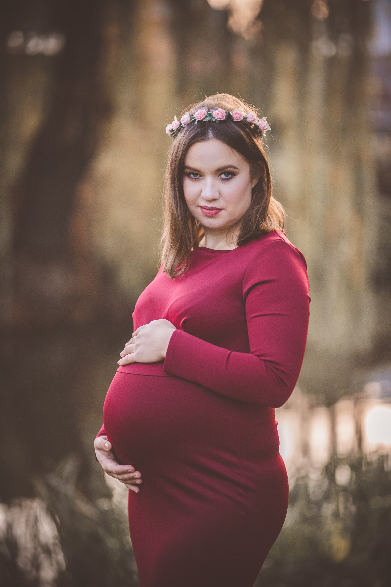 Kobieta w ciąży w bordowej sukience, z wiankiem z róż na głowie, pozuje w plenerze, trzymając ręce na brzuchu.
