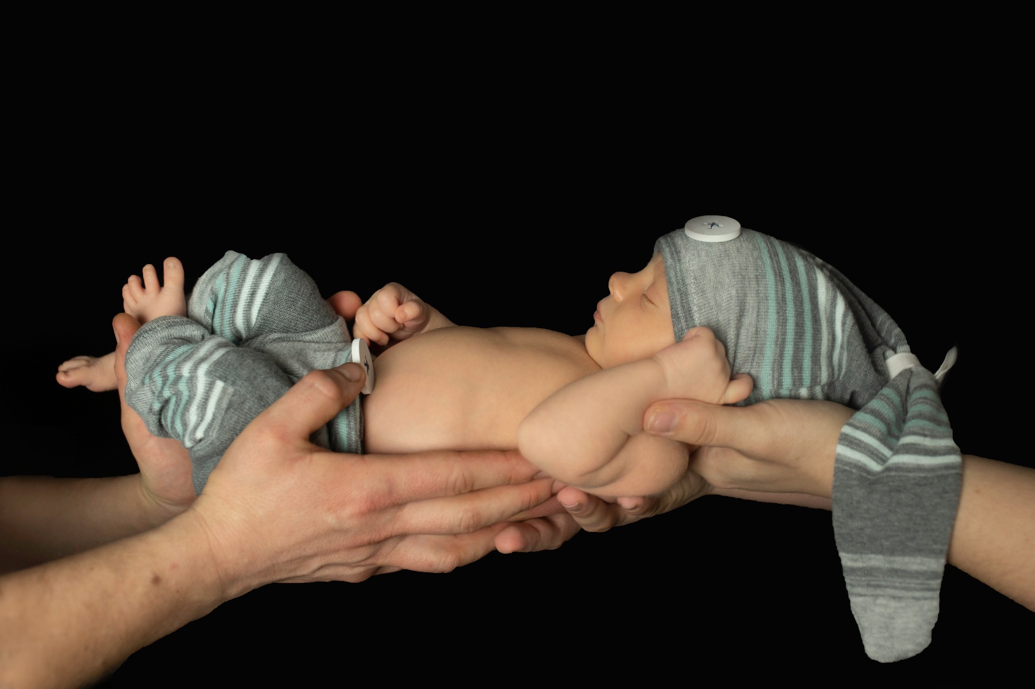 Noworodek w szaro-turkusowym, prążkowanym ubranku i czapeczce z guzikiem, trzymany w dłoniach na czarnym tle.