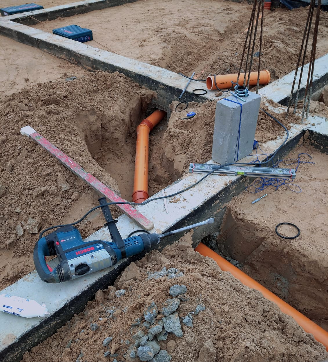 Pomarańczowe rury kanalizacyjne w wykopie fundamentowym, widoczny młot udarowy Bosch, poziomica i bloczek betonowy.