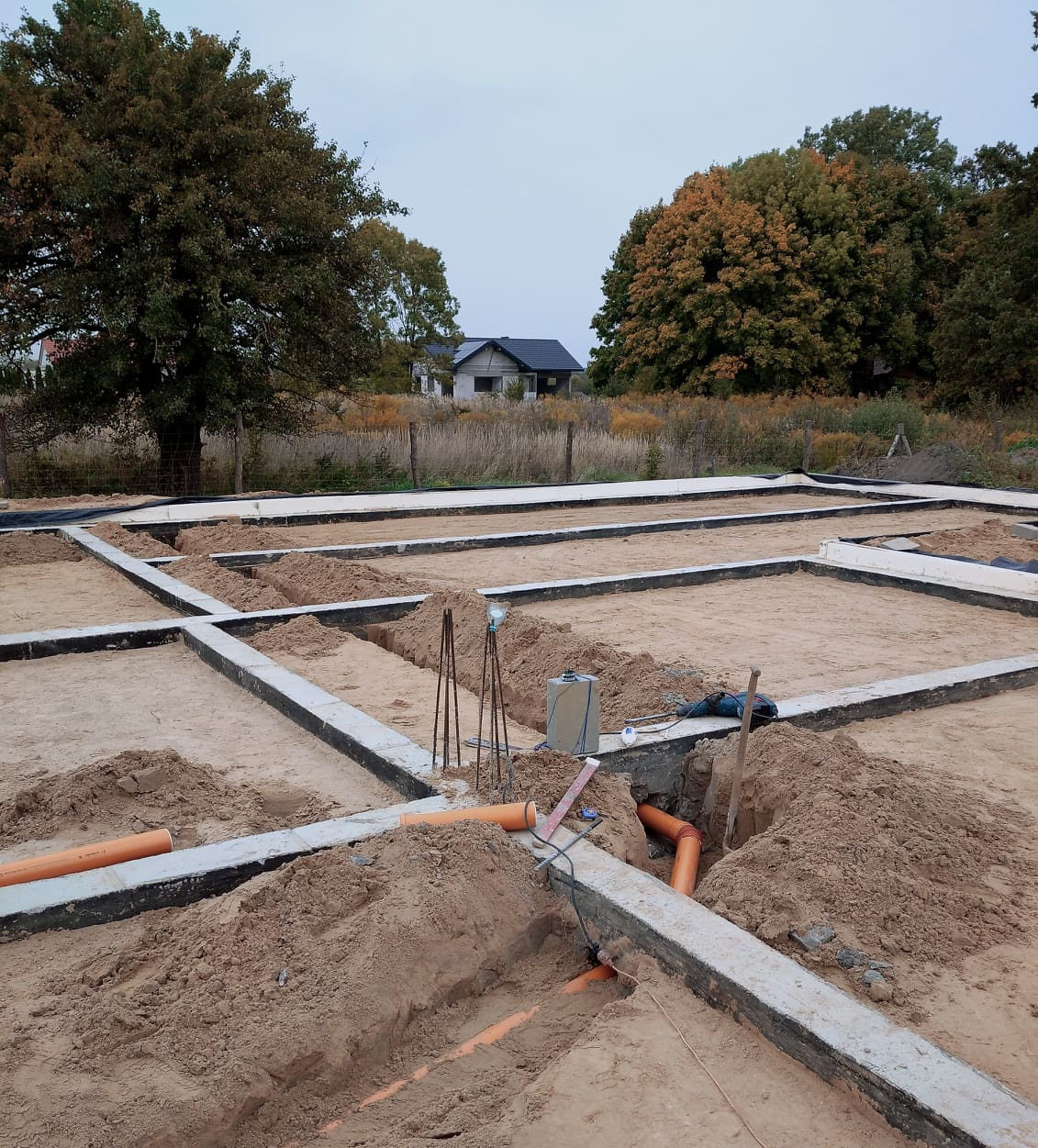 Fundamenty budynku w trakcie budowy z widocznymi rurami kanalizacyjnymi w wykopach, teren budowy z piaskiem i widokiem na dom w oddali.