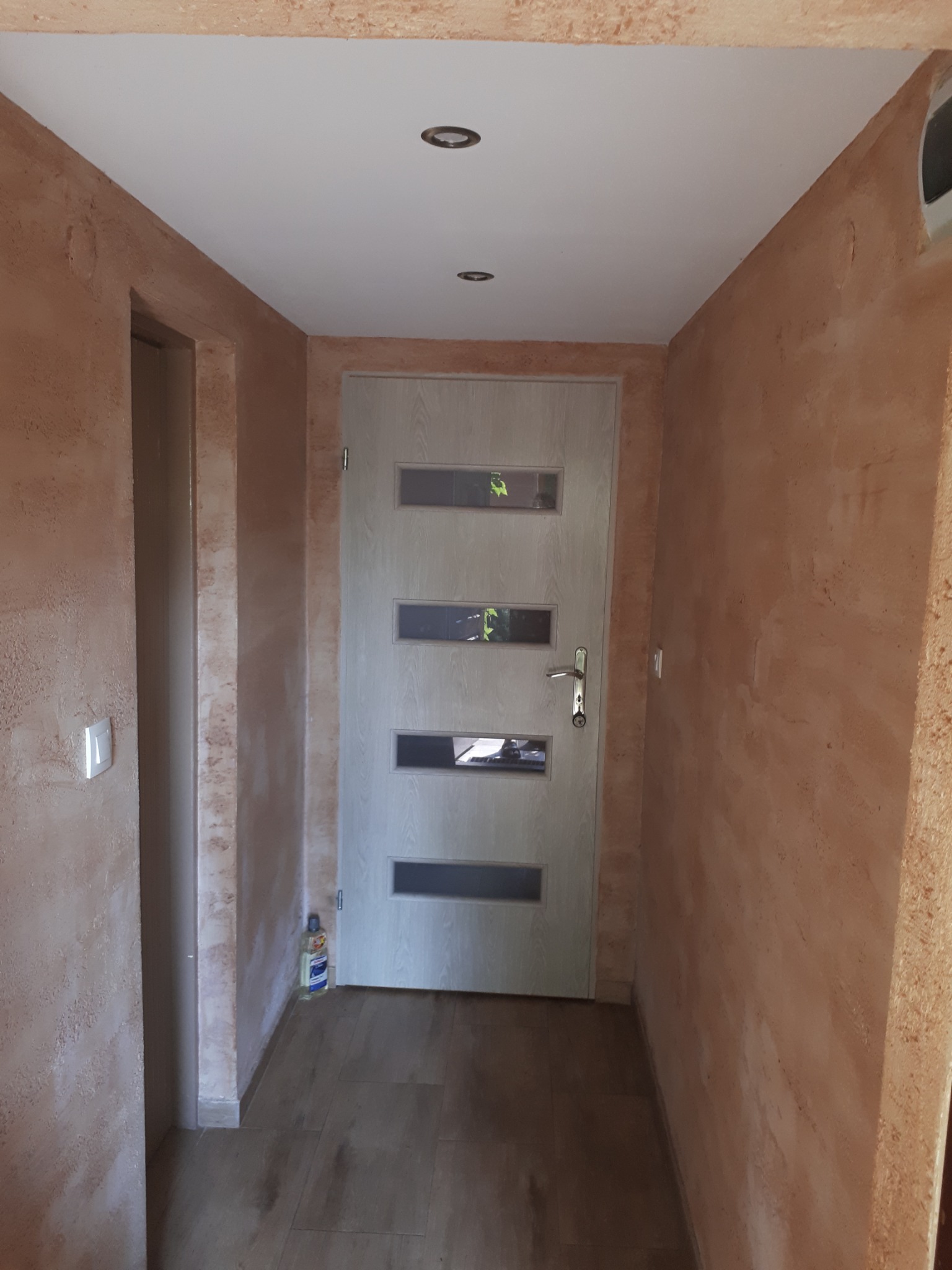 Korytarz po remoncie z dekoracyjną strukturą na ścianach w odcieniach beżu, drewnopodobne panele na podłodze, białe drzwi z czterema przeszkleniami i punktowe oświetlenie sufitowe.