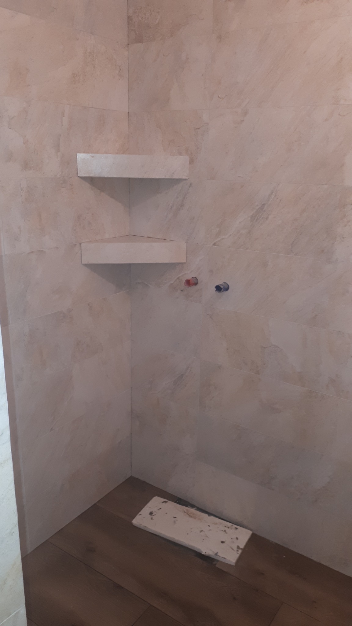 Narożna kabina prysznicowa z beżowymi płytkami imitującymi kamień, dwiema półkami narożnymi i widocznymi przyłączami wodnymi, na podłodze panele imitujące drewno, przy podłodze leży płytka