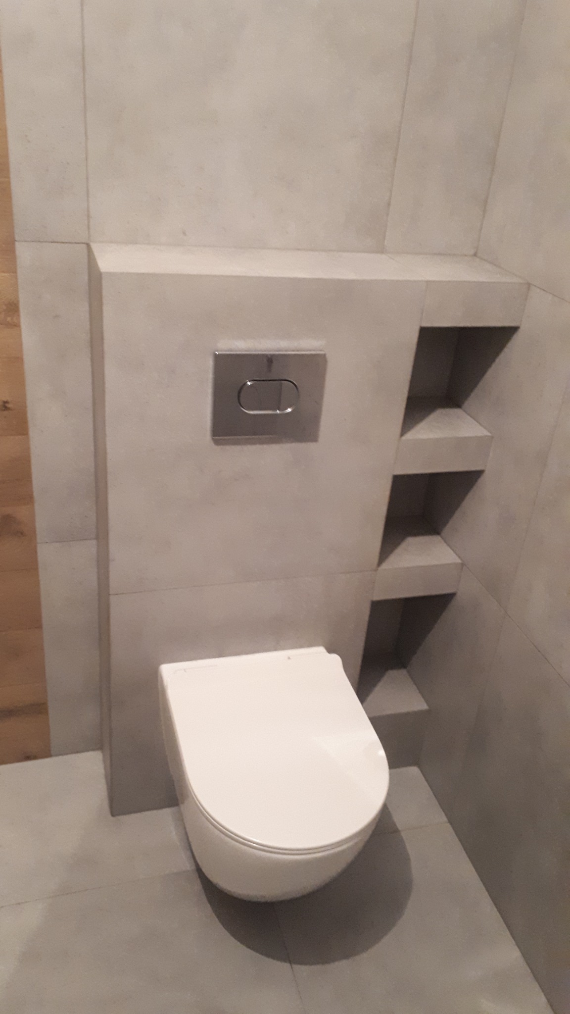 Nowoczesna toaleta podwieszana z szarą zabudową z półkami wnękowymi i chromowanym przyciskiem spłukującym, na szarych płytkach podłogowych i ściennych.