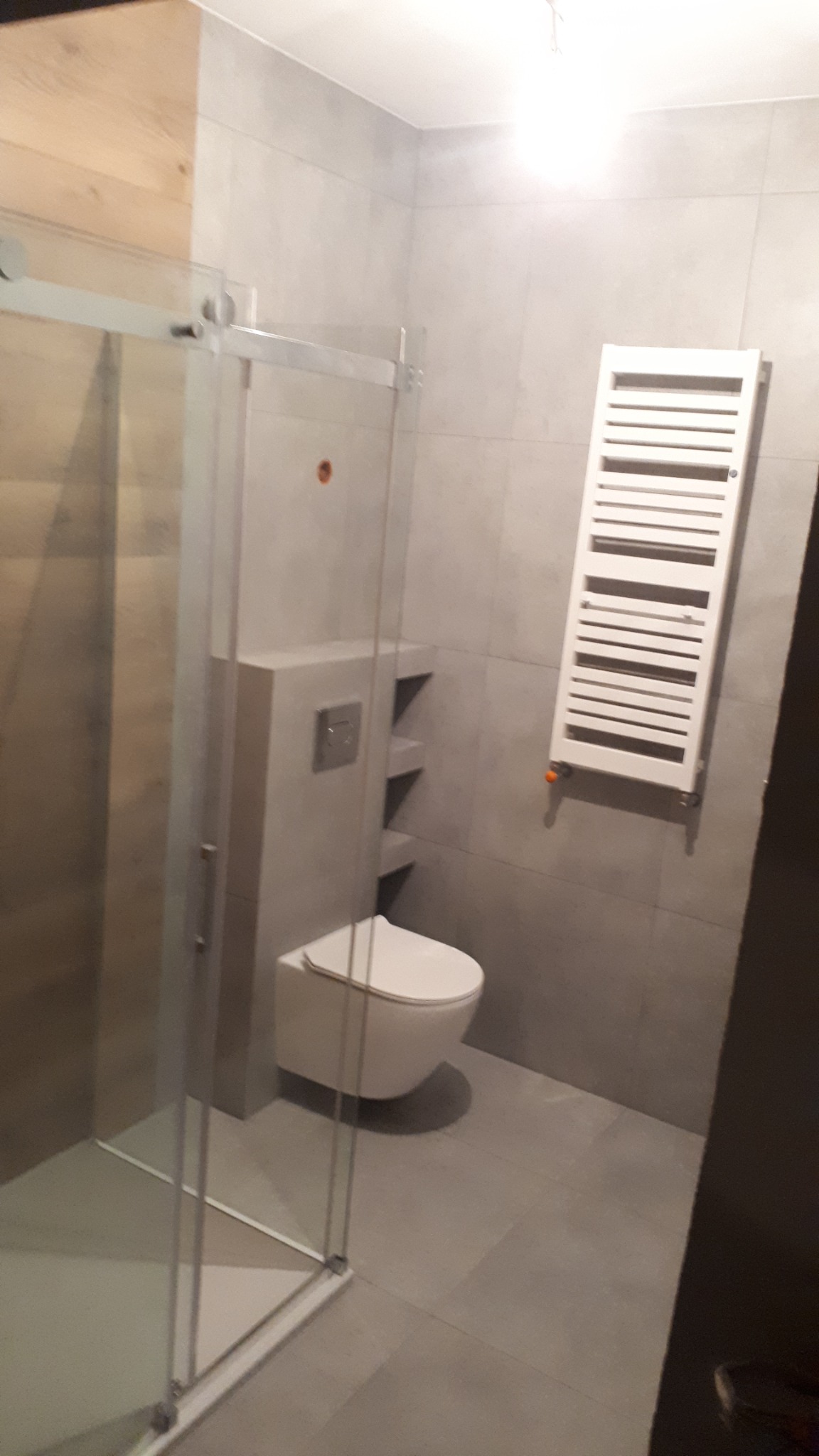 Nowoczesna łazienka z kabiną prysznicową typu walk-in, podwieszaną toaletą ze spłuczką w ścianie, grzejnikiem drabinkowym i szarą płytką ceramiczną.