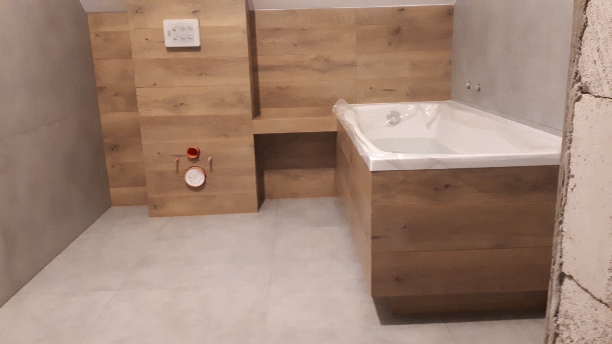 Wanna obudowana drewnopodobnymi płytkami, zamontowana armatura, szare ściany i podłoga, widoczne przyłącza wodne i elektryczne w trakcie prac wykończeniowych łazienki.