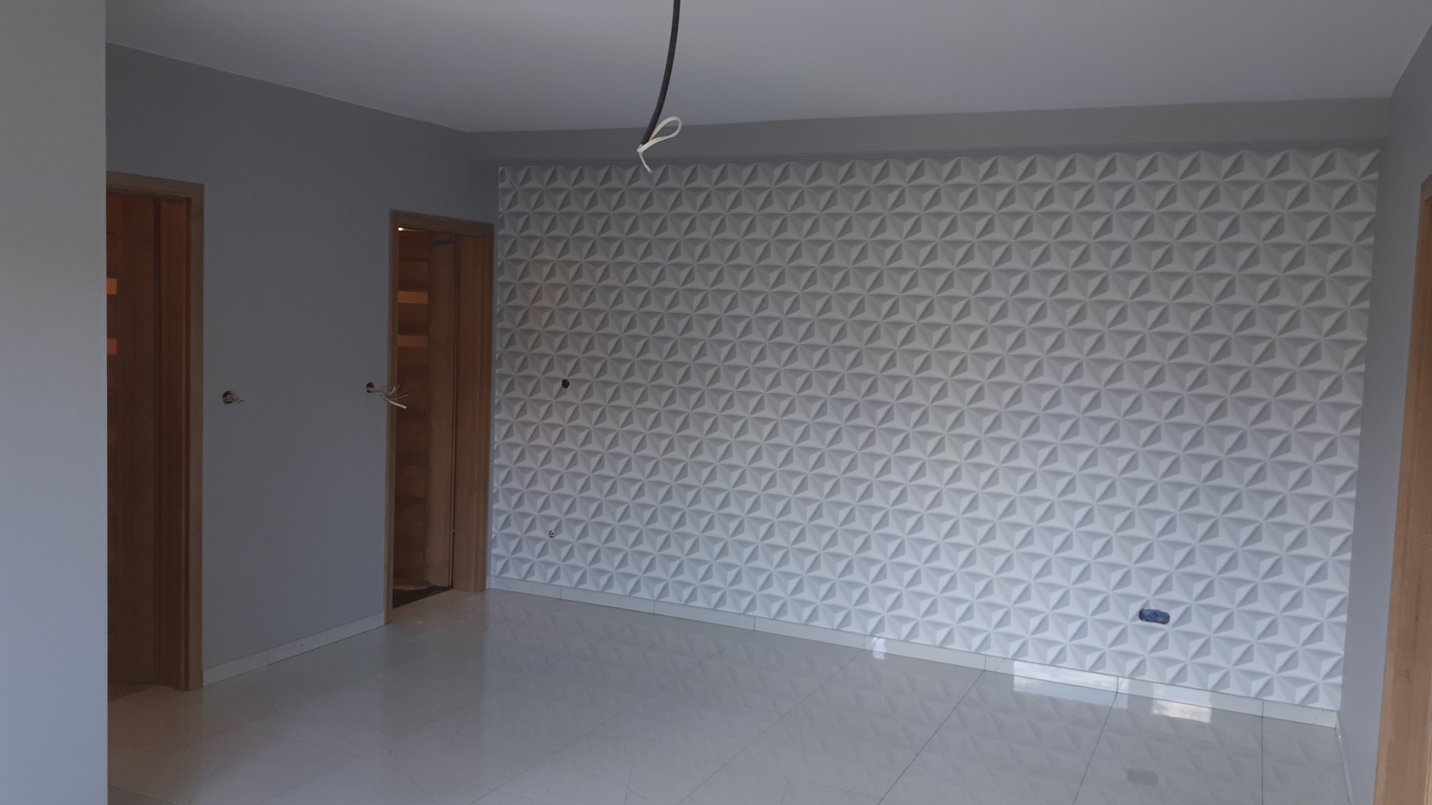 Pokój z białą trójwymiarową ścianą dekoracyjną, jasnymi płytkami podłogowymi i drewnianymi ościeżnicami drzwi. Widoczne wystające kable elektryczne.