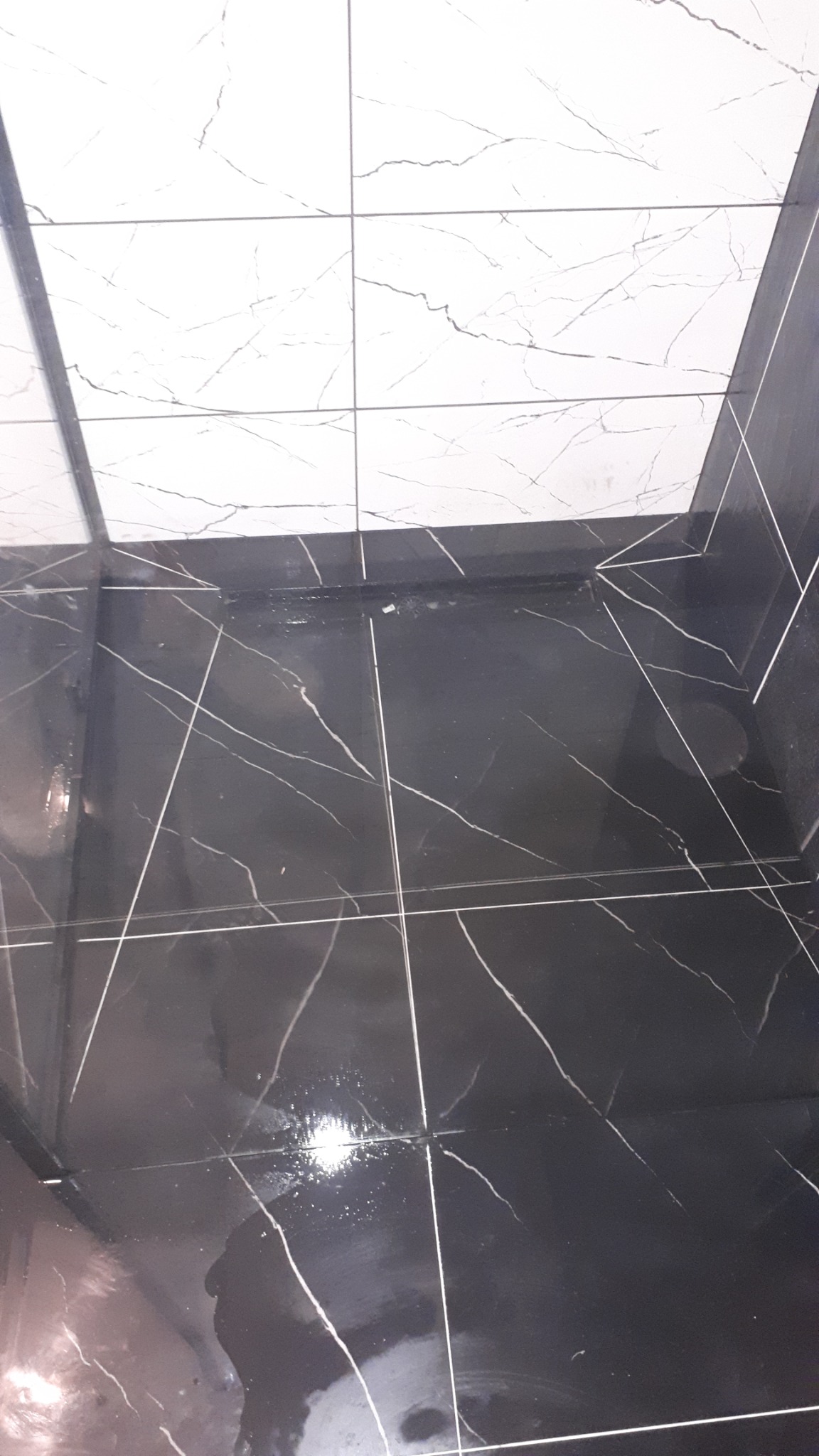 Biało-czarna glazura w łazience, widoczna podłoga i fragment ściany, z refleksami światła na ciemnych płytkach.