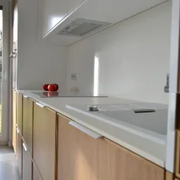 Nowoczesna kuchnia z drewnianymi frontami szafek dolnych i białymi szafkami górnymi, minimalistyczny design, trzy czerwone jabłka na płycie indukcyjnej, widok z boku, jasne oświetlenie.