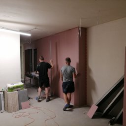Łukasz Szymkowiak - Dwóch mężczyzn montuje różowe płyty gipsowo-kartonowe na ścianie w pomieszczeniu w trakcie prac wykończeniowych, widoczne narzędzia, materiały budowlane i przewody elektryczne zwisające z sufitu.