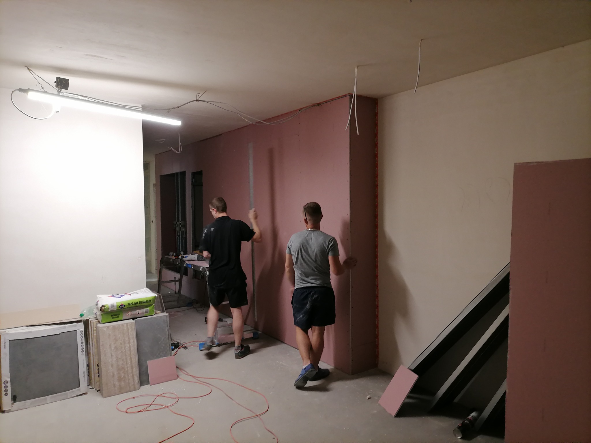 Dwóch mężczyzn montuje różowe płyty gipsowo-kartonowe na ścianie w pomieszczeniu w trakcie prac wykończeniowych, widoczne narzędzia, materiały budowlane i przewody elektryczne zwisające z sufitu.