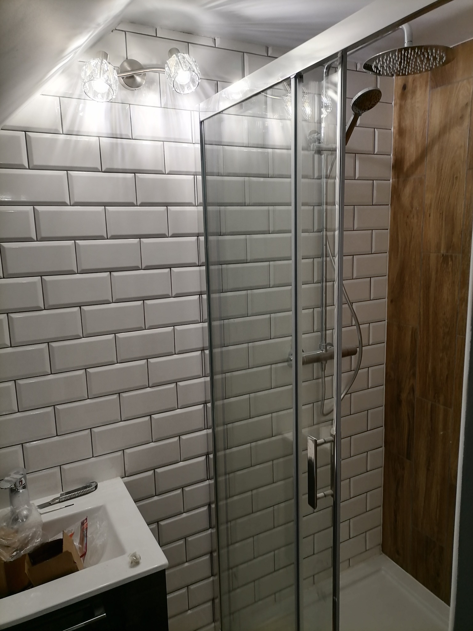 Nowoczesna kabina prysznicowa z deszczownicą, obudowana białymi, prostokątnymi płytkami ceramicznymi typu metro i panelami imitującymi drewno, oświetlona kryształowymi kinkietami.