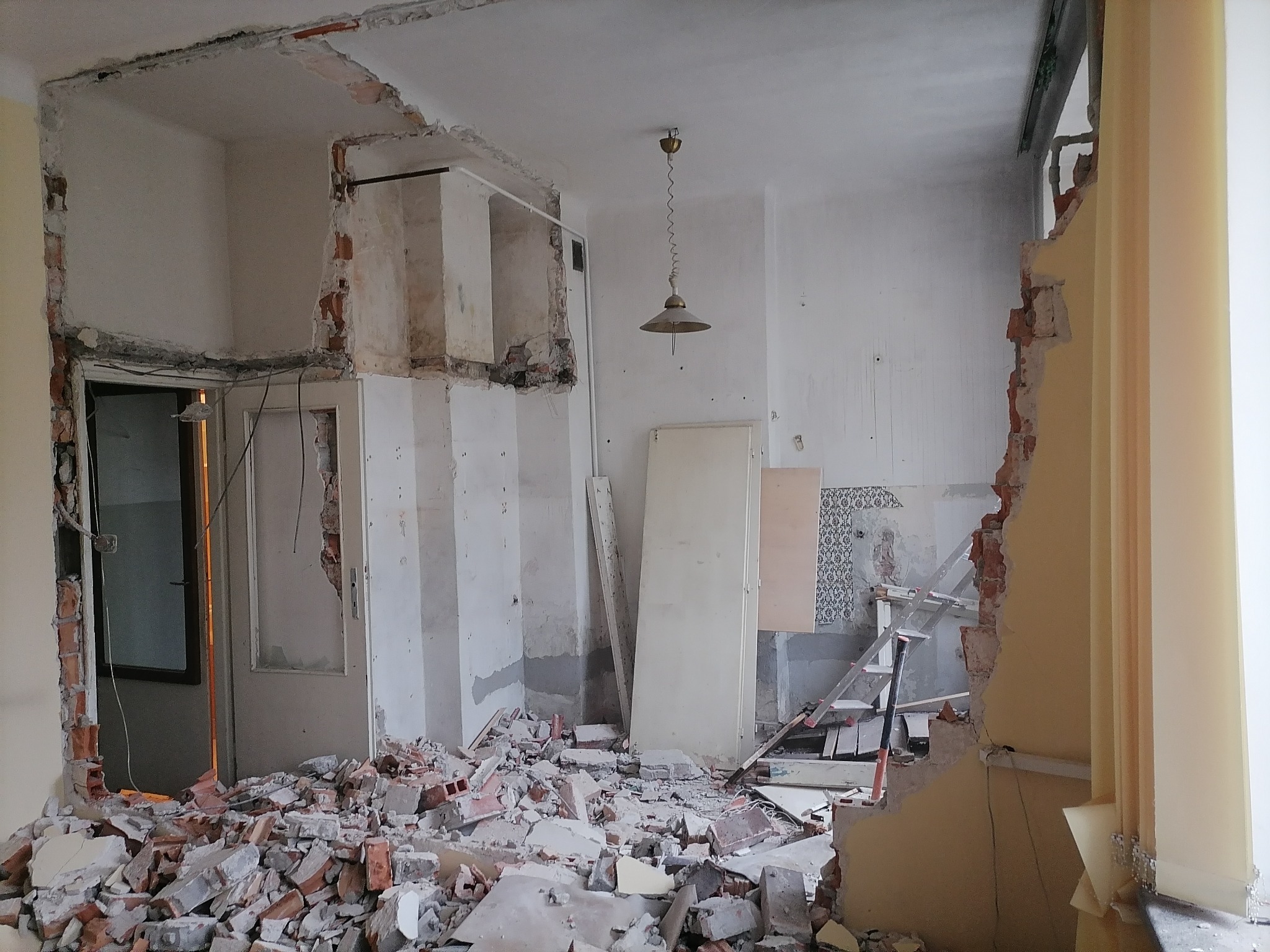 Pokój w trakcie demontażu: gruz ceglany i fragmenty ścian po wyburzeniach, widoczna instalacja elektryczna i stara lampa na suficie, zarys dawnej aranżacji wnętrza.