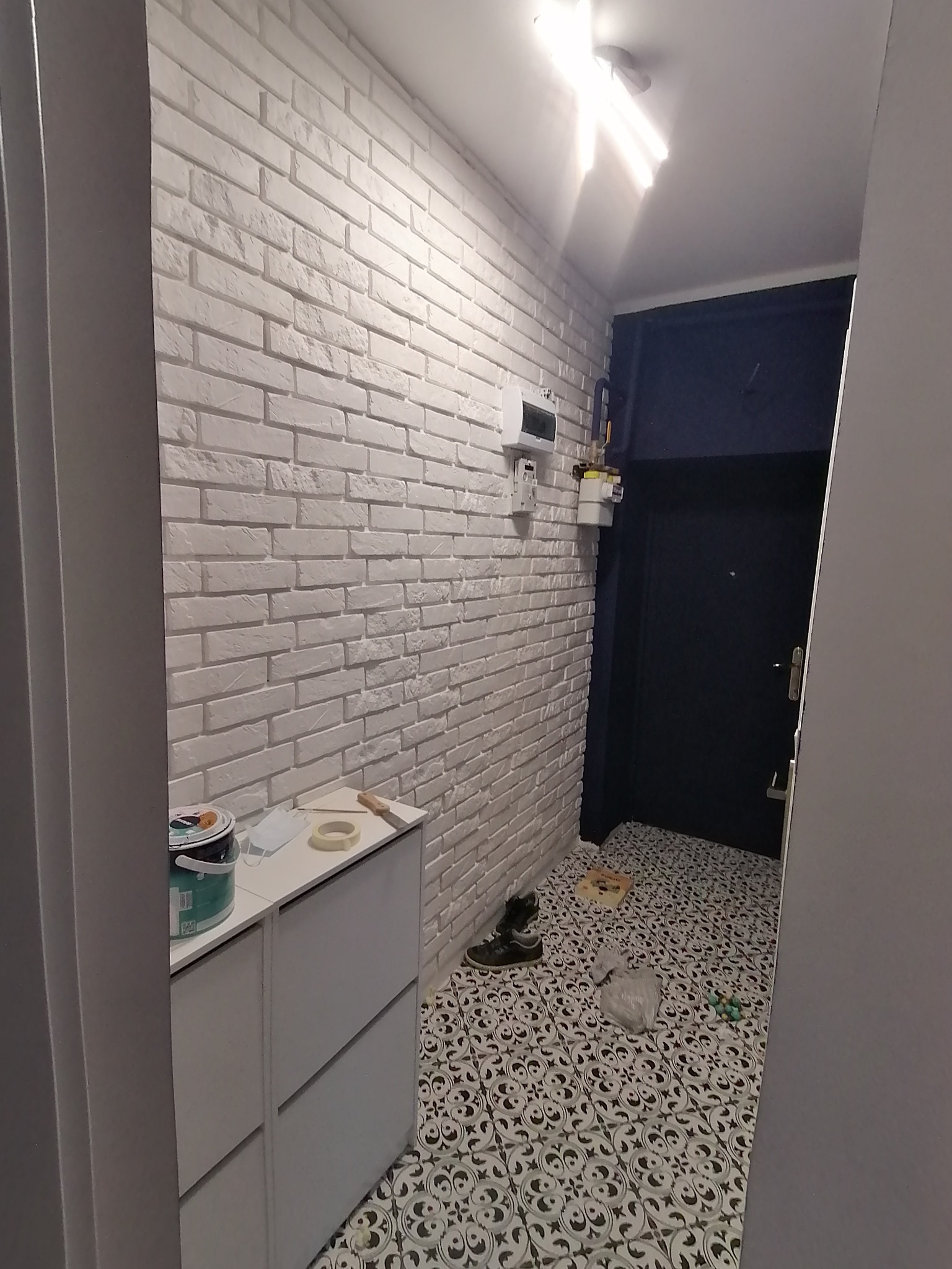 Korytarz w trakcie prac wykończeniowych, ściana wyłożona białą cegłą dekoracyjną, widoczne narzędzia malarskie i wiadro z farbą na szafce, podłoga wyłożona wzorzystymi płytkami.