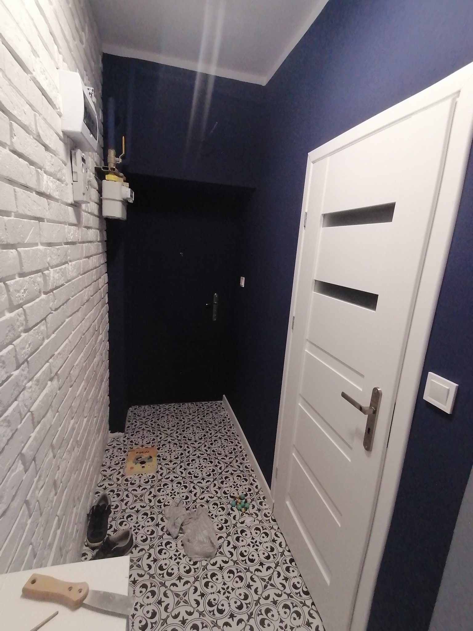 Korytarz po remoncie: biała cegła na ścianie, czarno-biała wzorzysta podłoga, białe drzwi z poziomymi przeszkleniami i granatowa ściana, widoczny licznik gazu i narzędzie szpachlarskie.