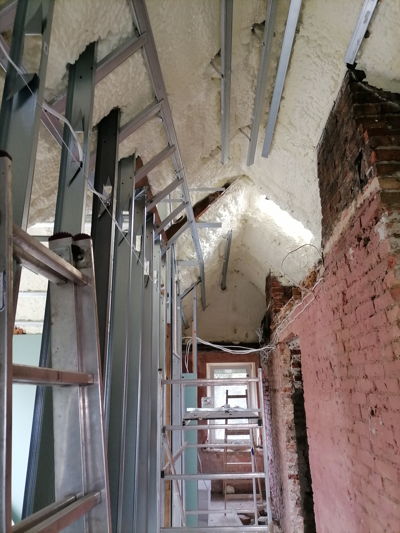 Montaż metalowej konstrukcji pod płytę karton-gips na poddaszu z widoczną izolacją piankową i fragmentem ceglanej ściany w trakcie prac wykończeniowych.